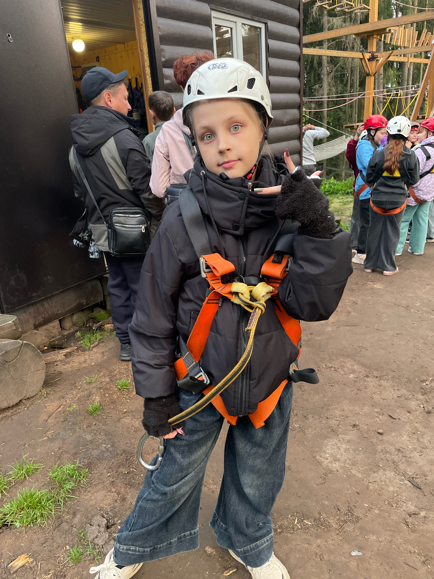 A young girl with blonde hair, wearing a white helmet, black jacket, and blue jeans, stands in the foreground of an outdoor scene. She is looking at the camera with her right hand raised in a "peace" sign. She is wearing a harness with orange straps and a metal carabiner. Other people, also wearing helmets and harnesses, are visible in the background, as well as a wooden structure and trees. The ground is dirt and grass. The image appears to have been taken in Pervomaysky, Russia.