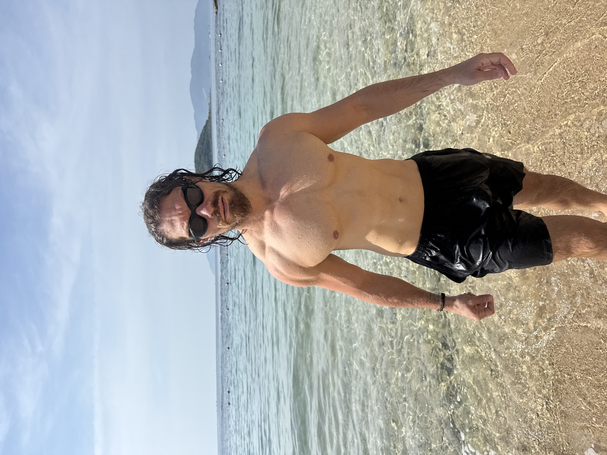 A man with wet, dark, curly hair and a beard, wearing black swim trunks and sunglasses, stands on a sandy beach. He is partially submerged in clear water. The ocean stretches out behind him, with several buoys visible in the distance. In the background, mountains rise against a pale blue sky with scattered clouds. The scene is located in the city of Bac Nha Trang Ward, Vietnam.