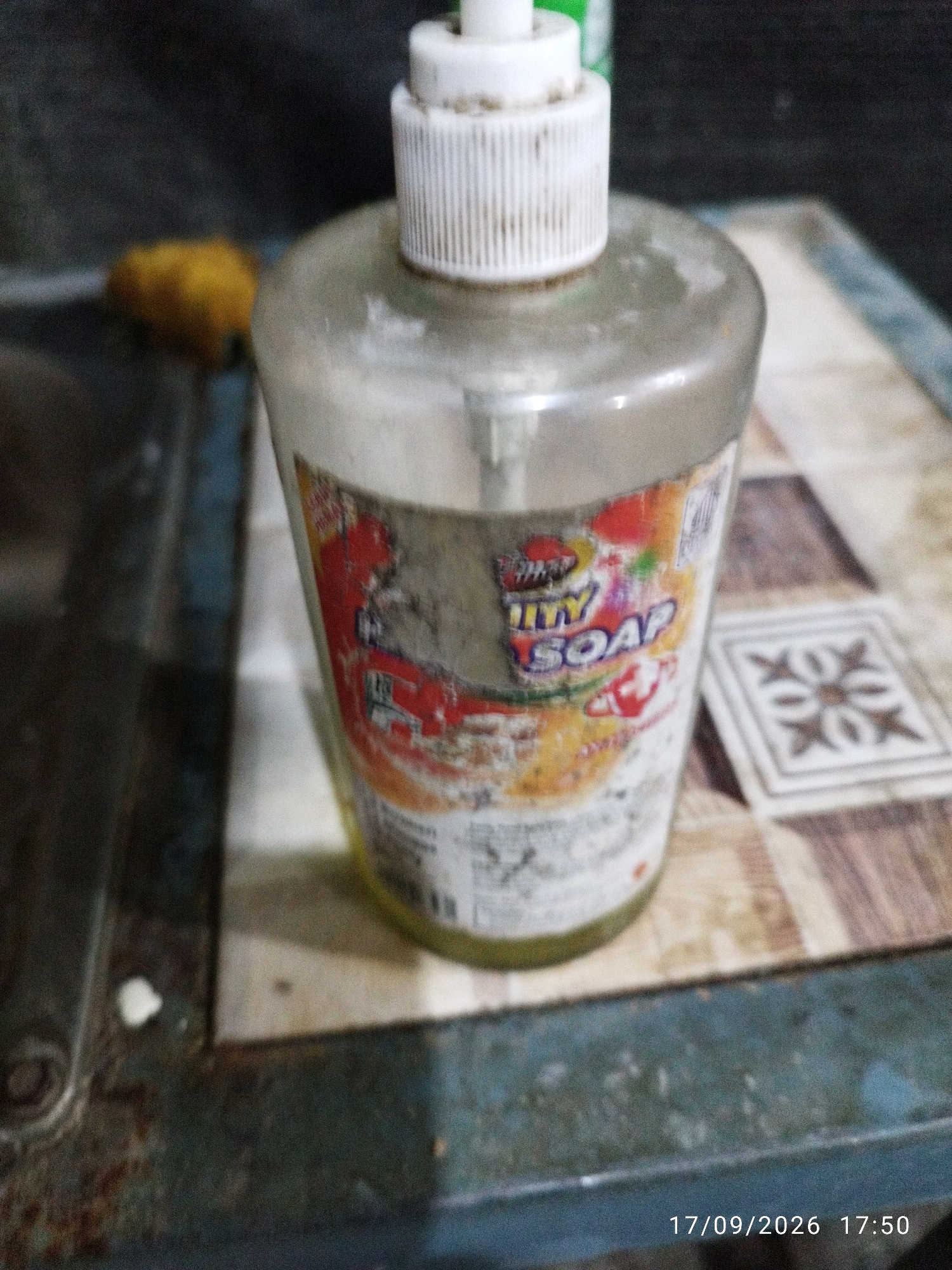 A clear plastic bottle of hand soap with a white pump dispenser is positioned on a tiled surface. The label on the bottle is partially obscured and appears to have red and yellow elements, with the text "ITY SOAP" visible. To the left of the soap bottle, a blurry yellow object is present. The background is dark and out of focus. The lower right corner of the image displays a date and time stamp: "17/09/2026 17:50". The scene appears to be indoors.