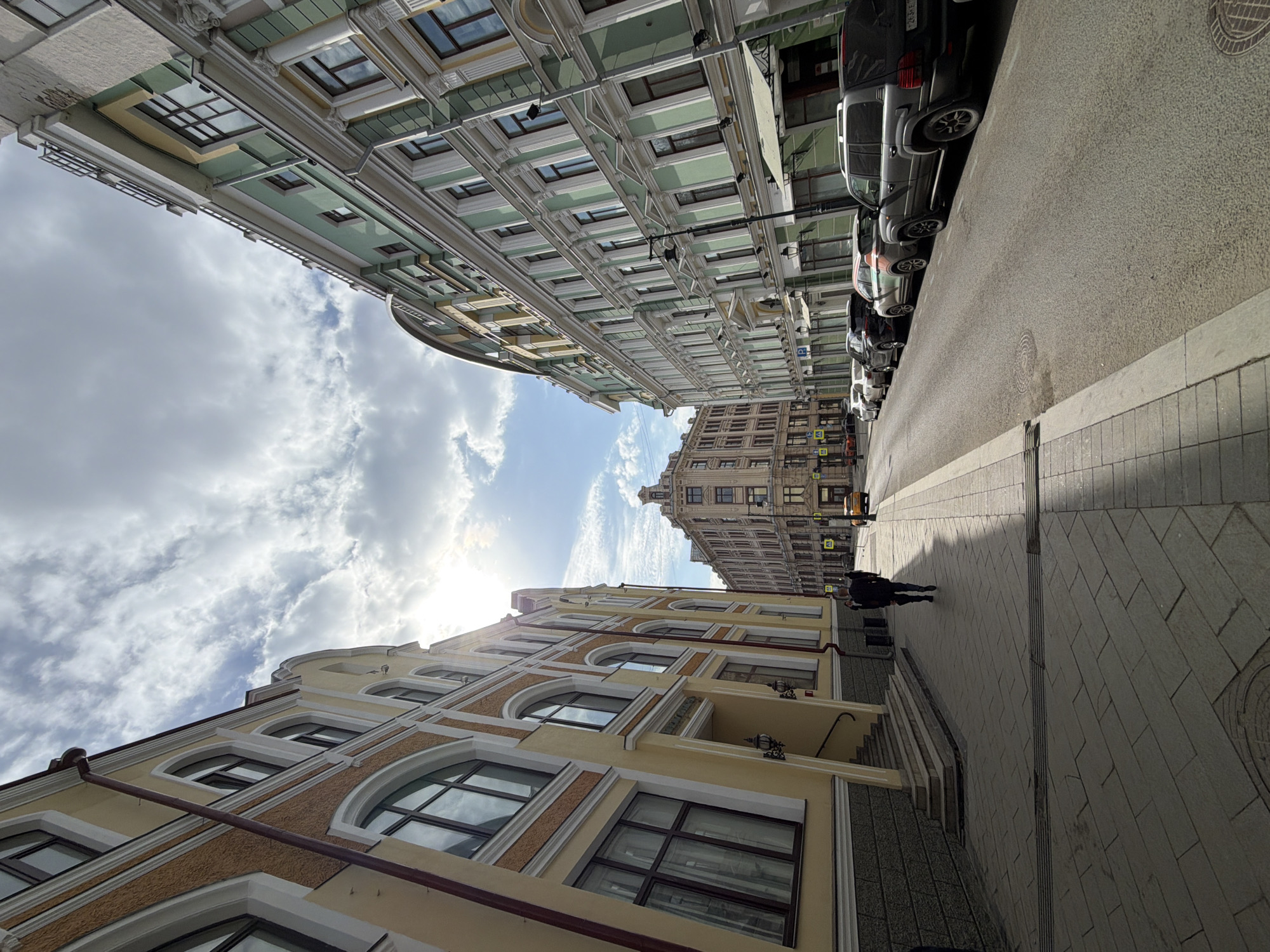 The image depicts a street scene in Moscow, Russia, characterized by tall buildings lining both sides of a narrow street. On the left side, a building is colored in yellow, while the other building is painted in green. Parked vehicles are seen along the right side of the street. The sky above is partly covered with clouds, letting sunlight through. A person is walking on the sidewalk.