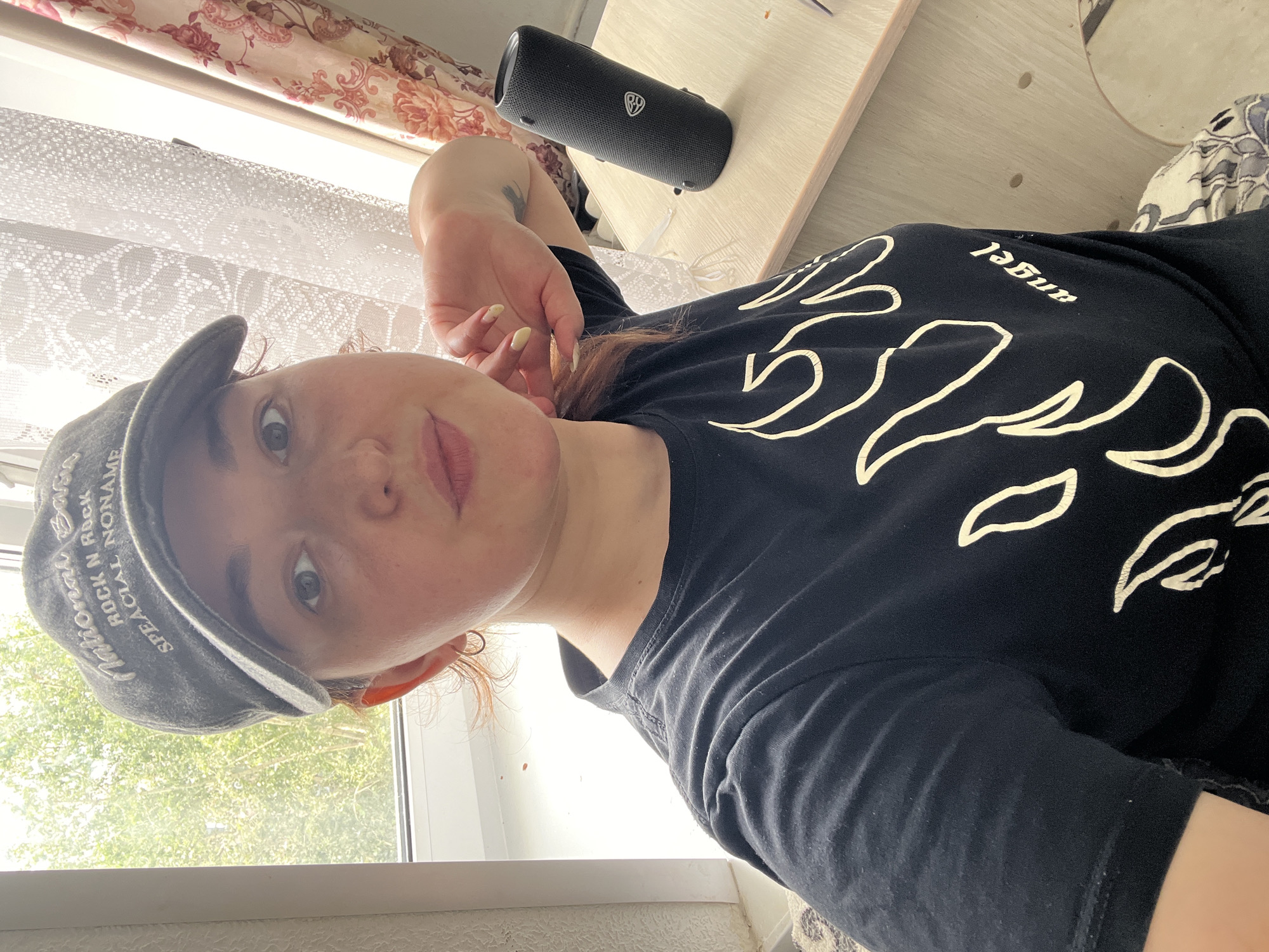 A young woman with auburn hair is captured in a selfie, wearing a dark baseball cap and a black t-shirt with a white flame design and the word "angel." Her right hand is resting on her chin, and her left arm is visible. A black portable speaker is positioned on a wooden surface to her right, with a floral patterned curtain and a white lace curtain visible in the background, suggesting an indoor setting. The scene appears to be a home environment, possibly near a window, given the natural light and the presence of curtains. There are no visible indicators that this scene is located in Chelyabinsk, Russia.