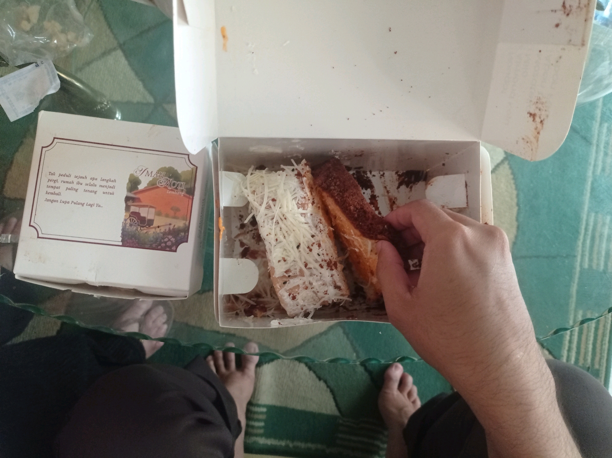 The image shows a person's hand reaching into a white cardboard box to pick up a piece of food. The box contains two pieces of food, one topped with shredded cheese and the other with a brown topping. There are crumbs scattered around the box. To the left of the box is another white cardboard container with text and an illustration. The scene appears to be set on a glass table over a carpet with a green and white pattern. In the foreground, the bare feet of at least two people are visible. The image context suggests this might be in Bandung City, Indonesia, as evidenced by the text on the container which appears to be in Indonesian.