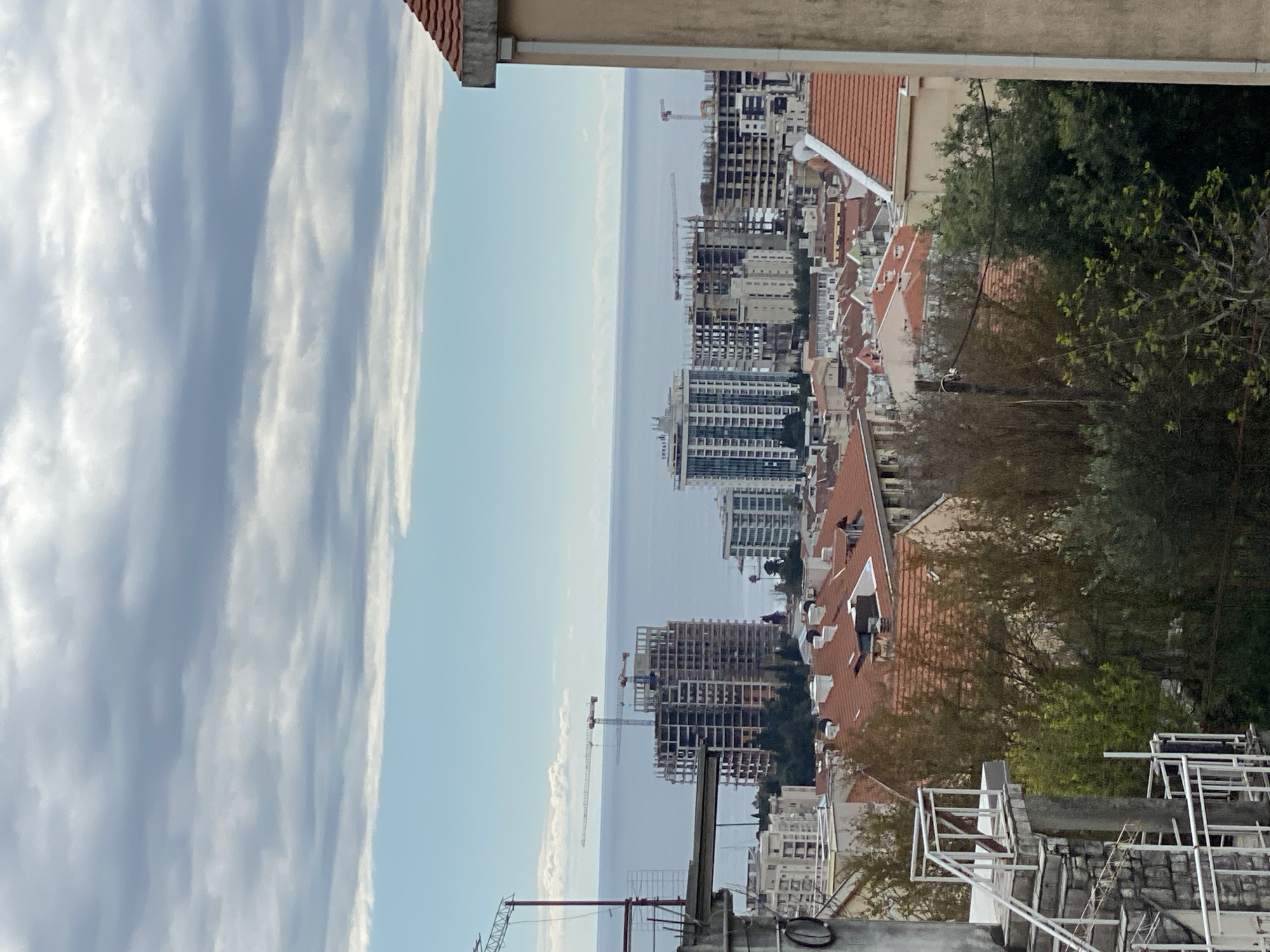 The image captures a cityscape with a view of the sea in Budva, Montenegro. Tall, modern buildings under construction are prominent, featuring cranes and skeletal structures. The older part of the city is characterized by buildings with red-tiled roofs. Trees provide green contrast. In the foreground, rooftop elements like railings and equipment are visible. The sky is filled with textured clouds.