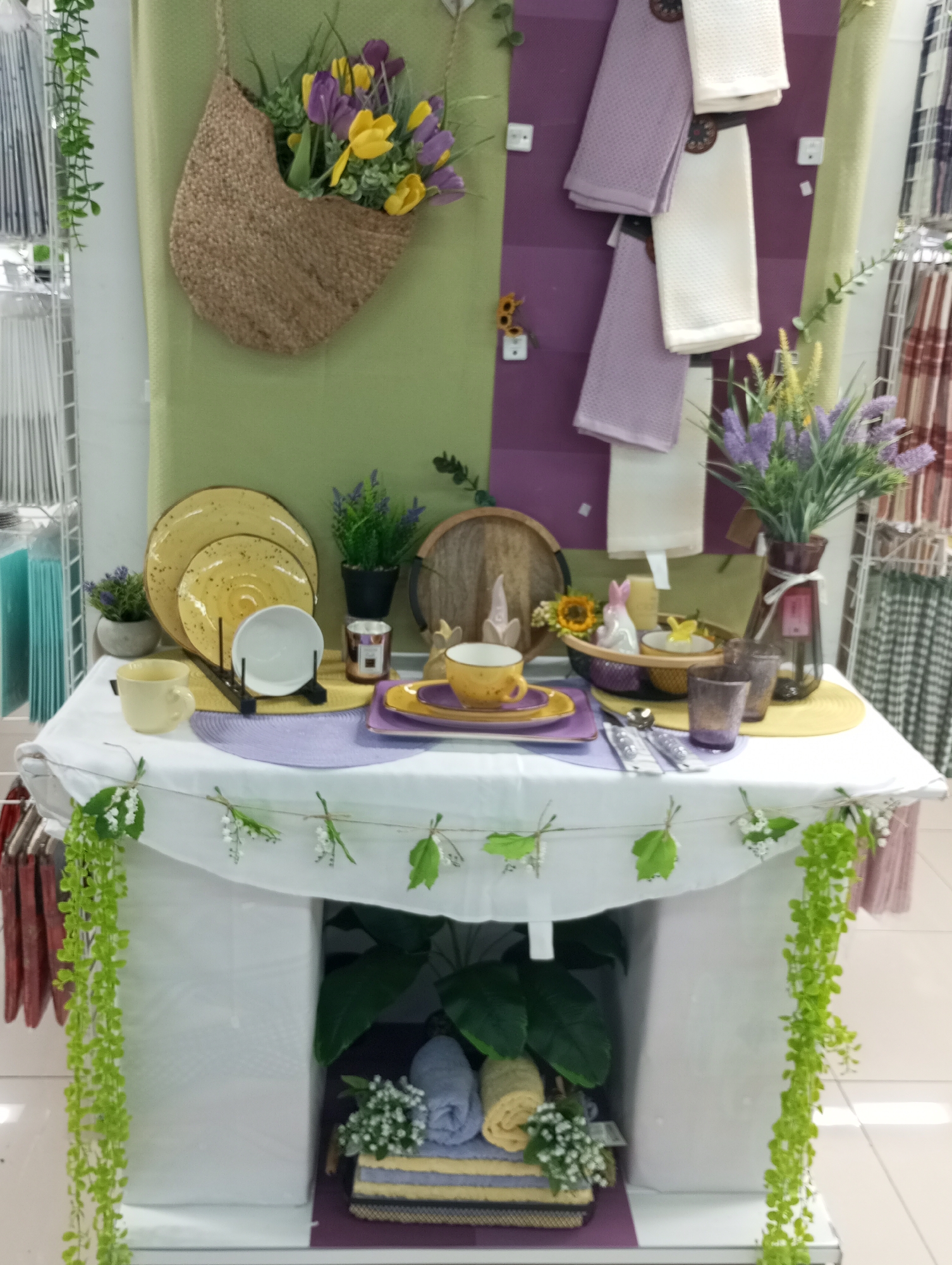 The image depicts a decorated display, likely in a retail setting. The backdrop is a dual-toned panel, green and purple, adorned with a hanging woven basket filled with artificial yellow and purple flowers. On the purple side are hanging white and light purple kitchen towels.
A white-clothed table stands in front of the backdrop, featuring a spring-themed tablescape. There are yellow plates, a white bowl on a stand, a yellow teacup and saucer, a wooden serving tray, and a basket containing a bunny figurine and small yellow flowers. A vase of artificial lavender sits toward the right, alongside two small purple glasses and silverware. Purple placemats are present beneath the dishes.
A garland of leaves and tiny white flowers decorates the edge of the tablecloth. Beneath the table, on a lower shelf, are rolled-up towels and artificial greenery, including potted plants.
The location is suggested to be Izhevsk, Russia, but there is no verifiable information within the image to confirm this.