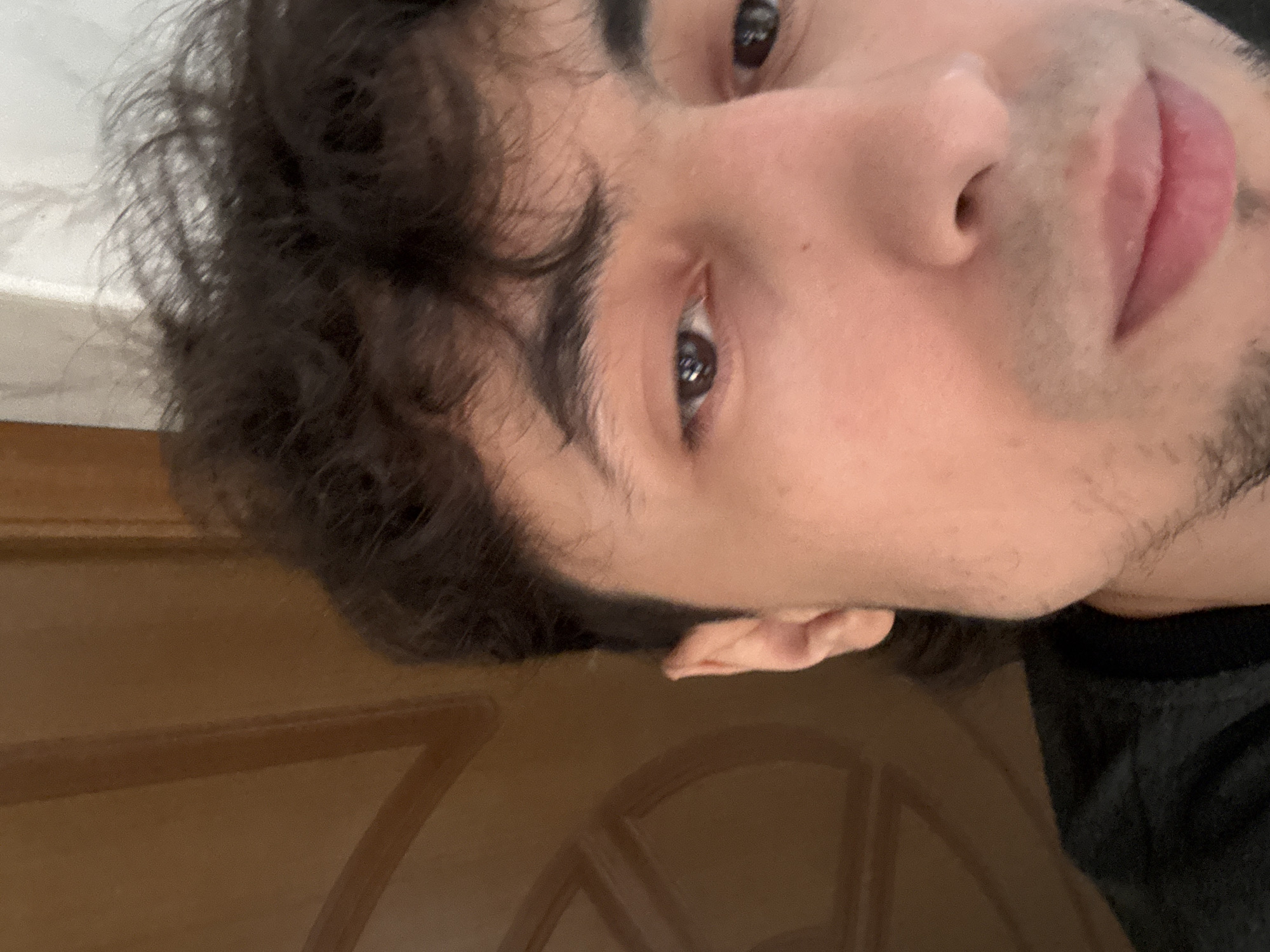 The image is a close-up, forward-facing, eye-level selfie of a young man's face and upper chest. He has dark, curly hair and is wearing a black t-shirt. The background is out of focus, showing what appears to be a wooden door with carved details on the left and a white curtain or wall on the right. The lighting is soft and appears to be from the front. The man's facial expression is neutral.