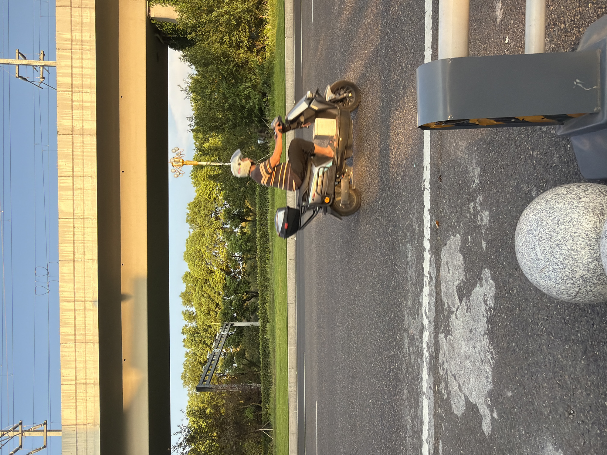 In Jiangjin, China, a person wearing a striped shirt and dark pants rides a light-colored scooter on an asphalt road. The scooter has a black storage box attached to the rear. The person is wearing a white helmet with a clear visor. The scene is set under an elevated concrete overpass, with utility poles and wires visible against a clear blue sky. Lush green trees line the side of the road beyond a grassy median. In the foreground, a gray and white speckled spherical object and a gray traffic barrier with yellow accents are visible. The lighting suggests it is either early morning or late afternoon, casting warm light and shadows.
