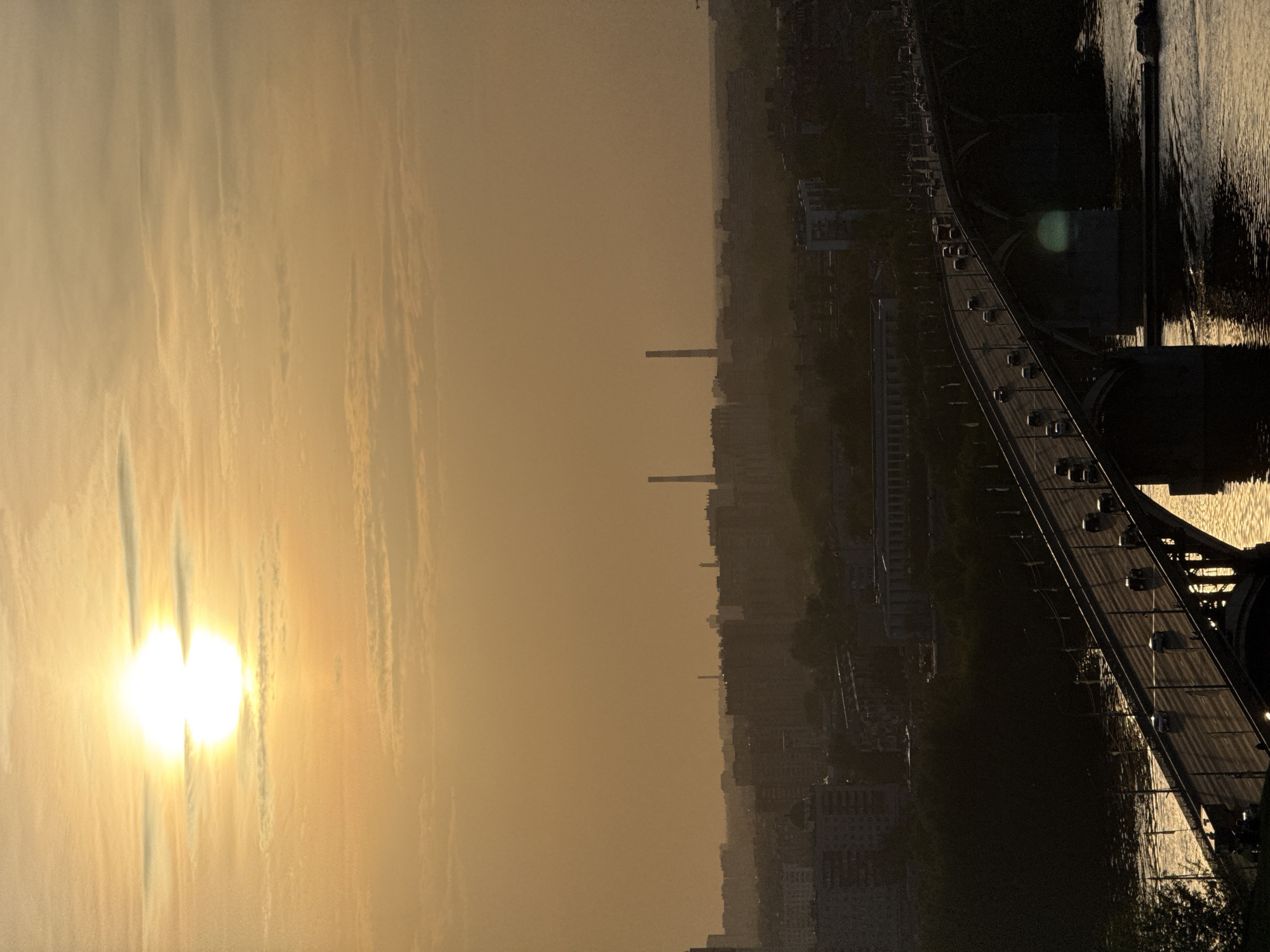 The image shows a sunset over the city of Nizhny Novgorod, Russia. The sun is low in the sky, casting a golden light over the city and the clouds. In the foreground, a bridge spans a river with light reflected on the water. Cars can be seen driving on the bridge. In the background, a dense urban landscape stretches into the distance, punctuated by tall chimneys or towers. The overall atmosphere is hazy, due to the setting sun and possible atmospheric conditions.