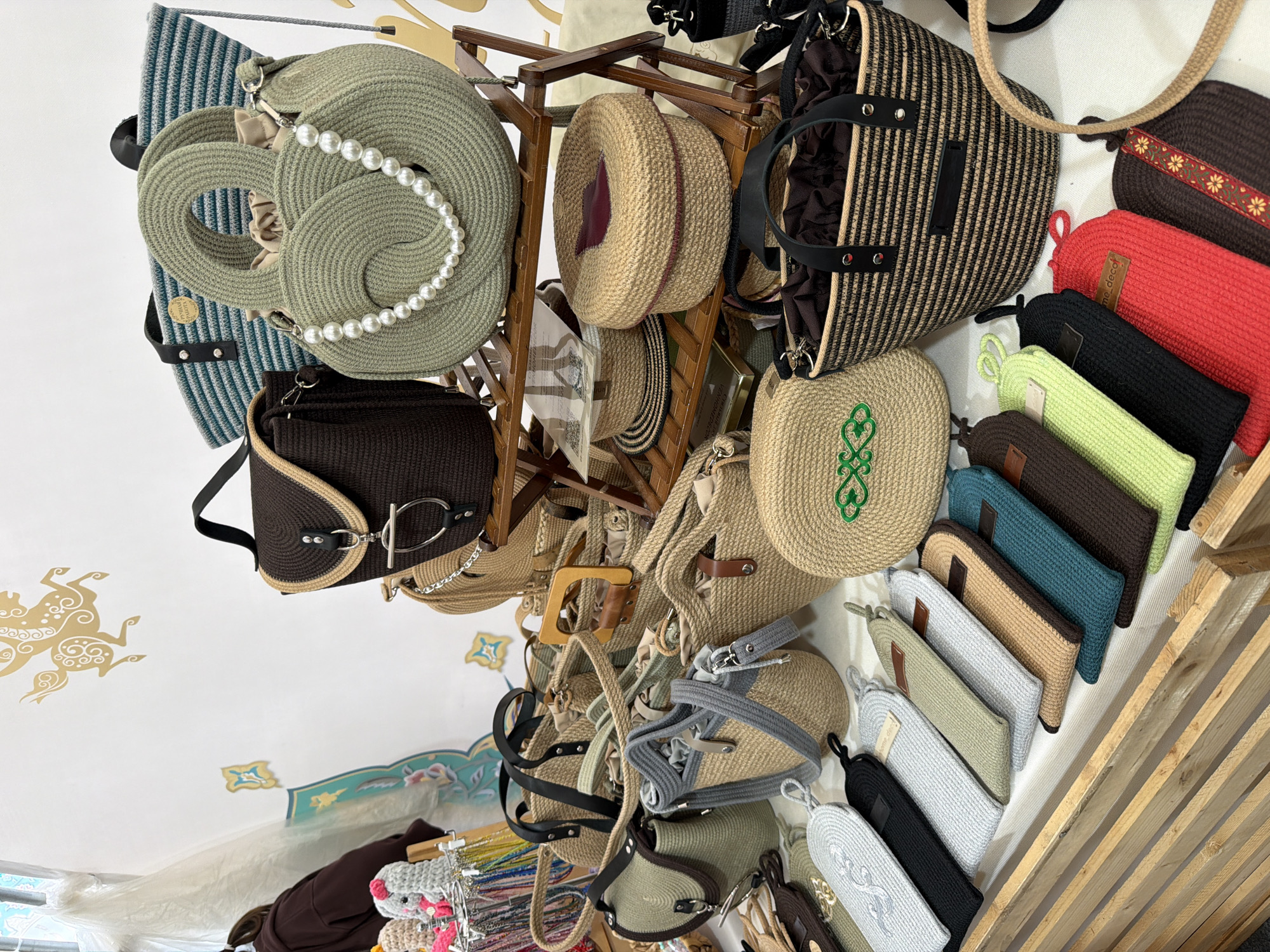An indoor market stall displays a variety of handmade woven bags and accessories. The items are primarily made of natural fibers like jute and straw, featuring earthy tones and some vibrant accents. Prominently displayed are several round and structured handbags, some adorned with pearls or decorative stitching. Below these, a collection of flat, pouch-like items in various colors are arranged in rows. A wooden shelf unit holds some of the bags, while others are placed on a white surface. The background shows a white wall with decorative golden motifs and a glimpse of a window with sheer curtains. A person in a brown hoodie is partially visible on the far left. The overall impression is one of a craft fair or artisan market.