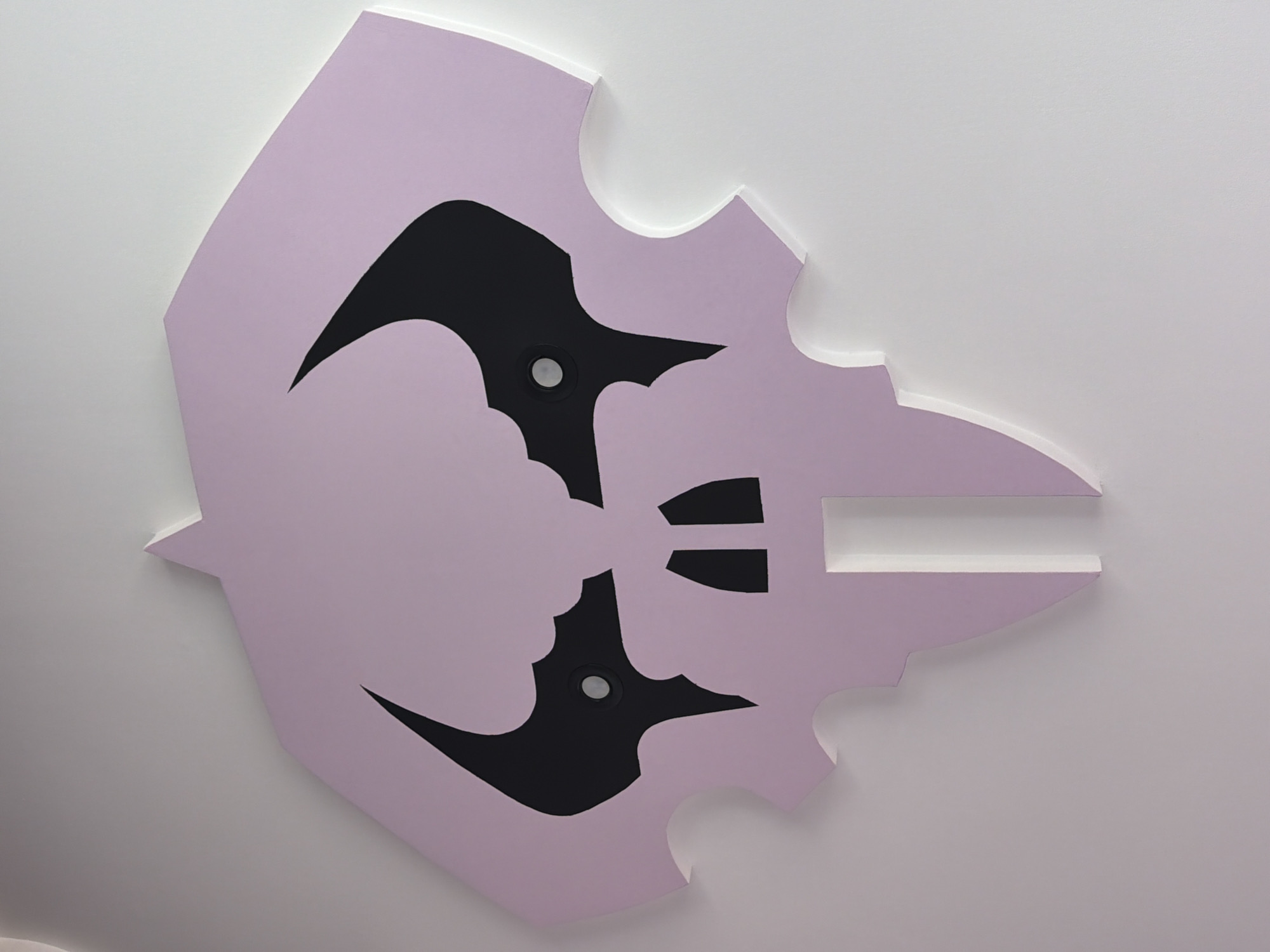 The image shows a close-up, top-down view of a light pink, stylized skull-shaped cutout mounted on a white wall. The cutout has large, black, almond-shaped eye sockets with small circular fixtures within them, and a stylized black mouth area forming two separate openings. The overall shape is angular with curved edges, reminiscent of a bat or a traditional skull motif. The background is a plain white wall, suggesting this is an indoor decorative piece. The location of Etropole, Bulgaria is not discernible from the visual elements present in the image.