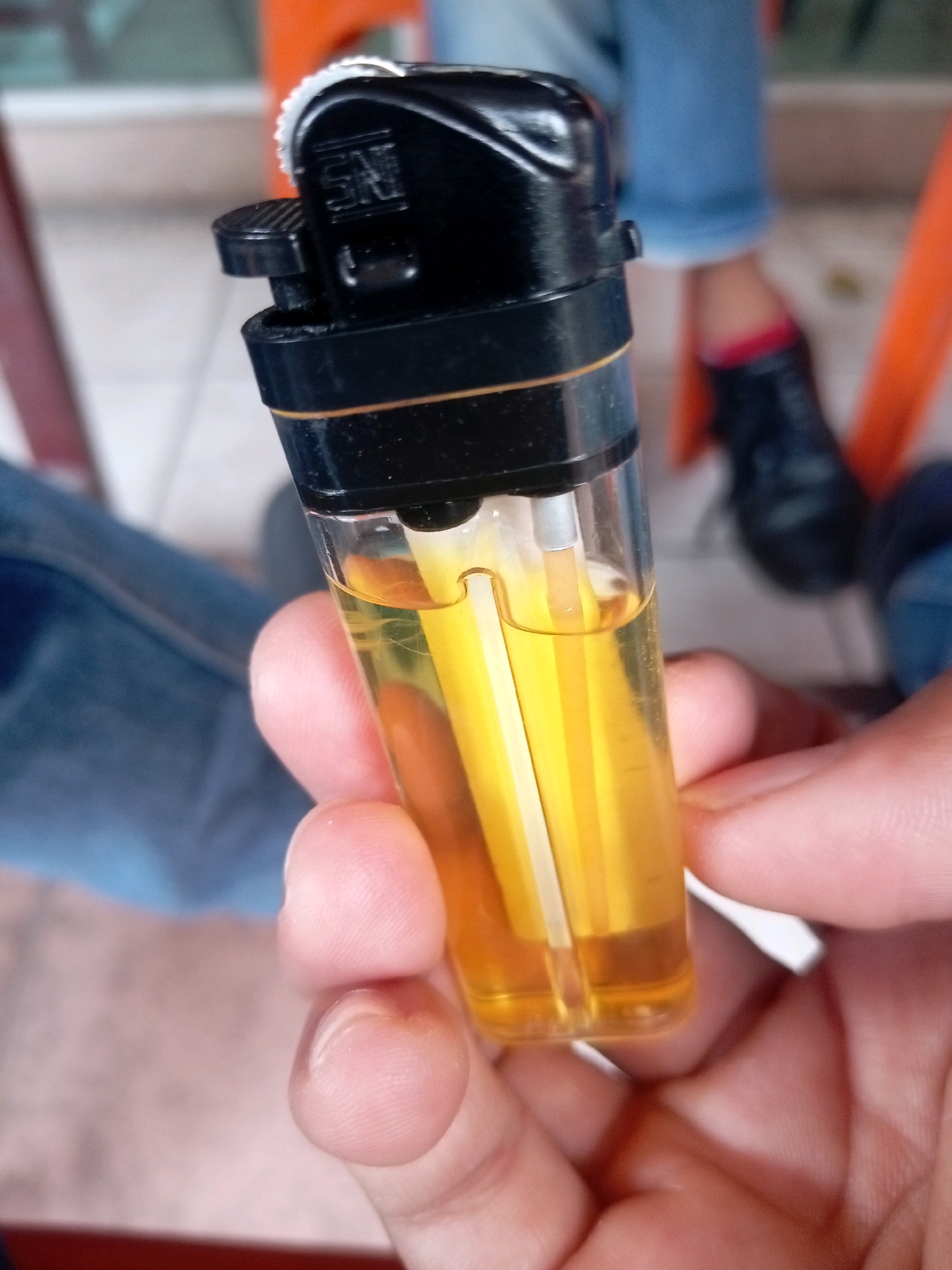 A close-up shot captures a transparent lighter filled with yellow fluid, held in a person's hand. The lighter's black top features a metallic scraping wheel. In the blurred background, a person wearing blue jeans and black shoes with red trim is visible. The scene appears to be indoors, possibly in a residential setting. The image does not provide specific details to pinpoint its location within Batam, Indonesia.