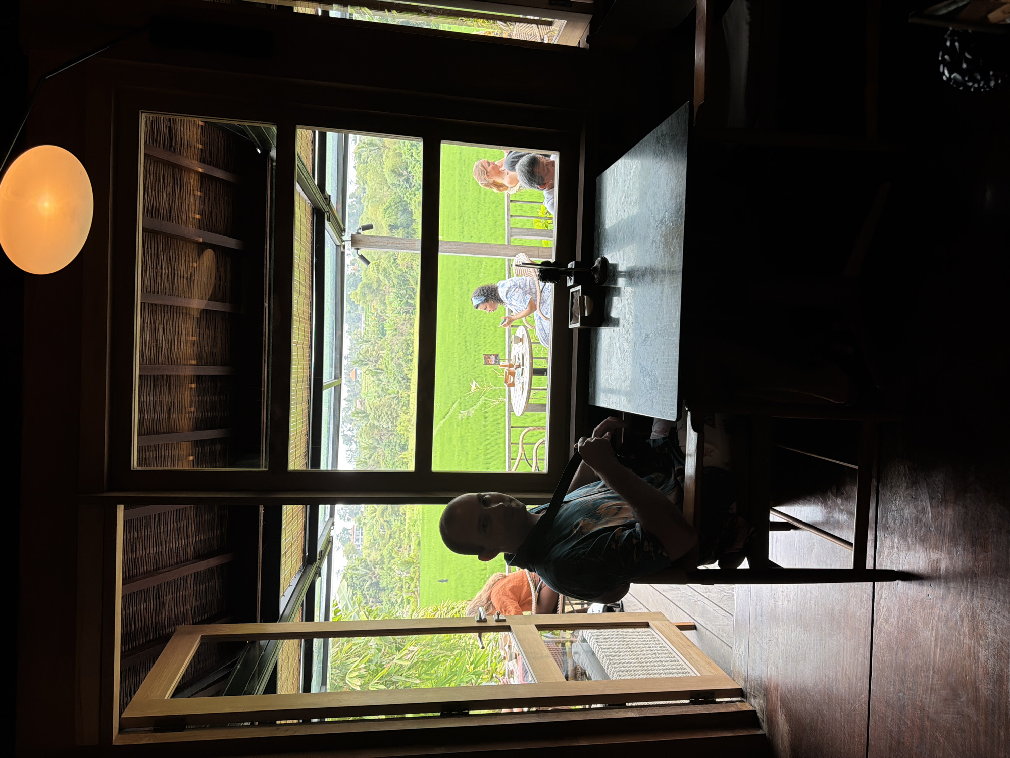 The image depicts the interior of a restaurant with a view of rice paddies in Sukawati, Indonesia. In the foreground, a man is seated at a dark-colored table, facing towards the right of the frame. He is wearing a patterned short-sleeved shirt and appears to be adjusting a strap or tie. A partially open wooden door is visible to the left, revealing a glimpse of the restaurant's outdoor seating area and lush greenery. Through the large windows, two women and one person whose gender is unclear are seated at tables on a patio overlooking vibrant green rice fields. The architecture features a thatched roof and wooden beams. An orange circular lamp hangs from the ceiling. The lighting suggests it might be daytime, with natural light filtering through the windows.