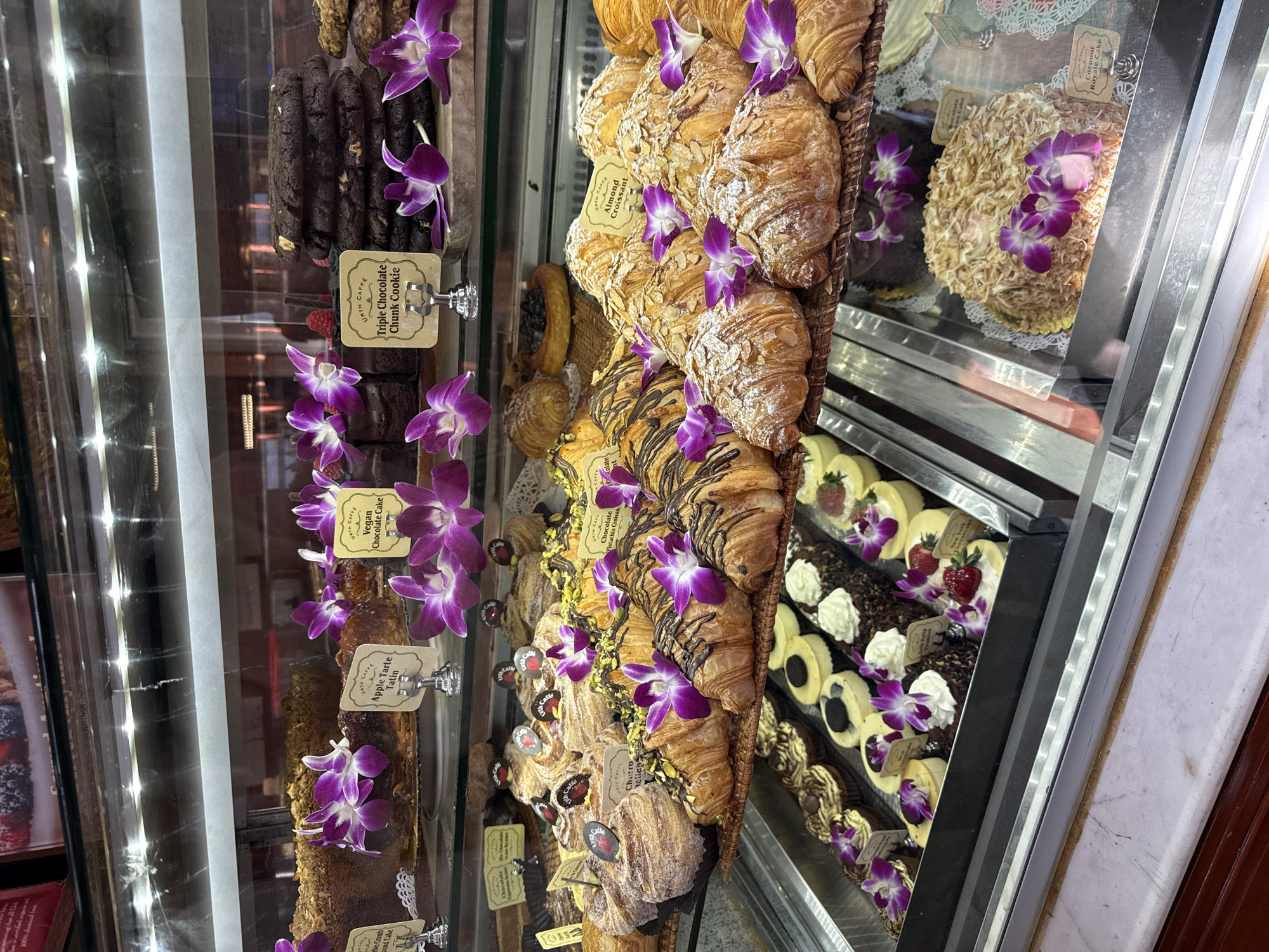 The image shows a display case filled with baked goods at Urth Caffé in Beverly Hills, United States of America. The case features items like almond croissants, chocolate pistachio croissants, triple chocolate chunk cookies, apple tarte tatin, vegan chocolate cake, and coconut royale cake. The items are decorated with purple orchids. Labels with the name "Urth Caffé" appear on each product.