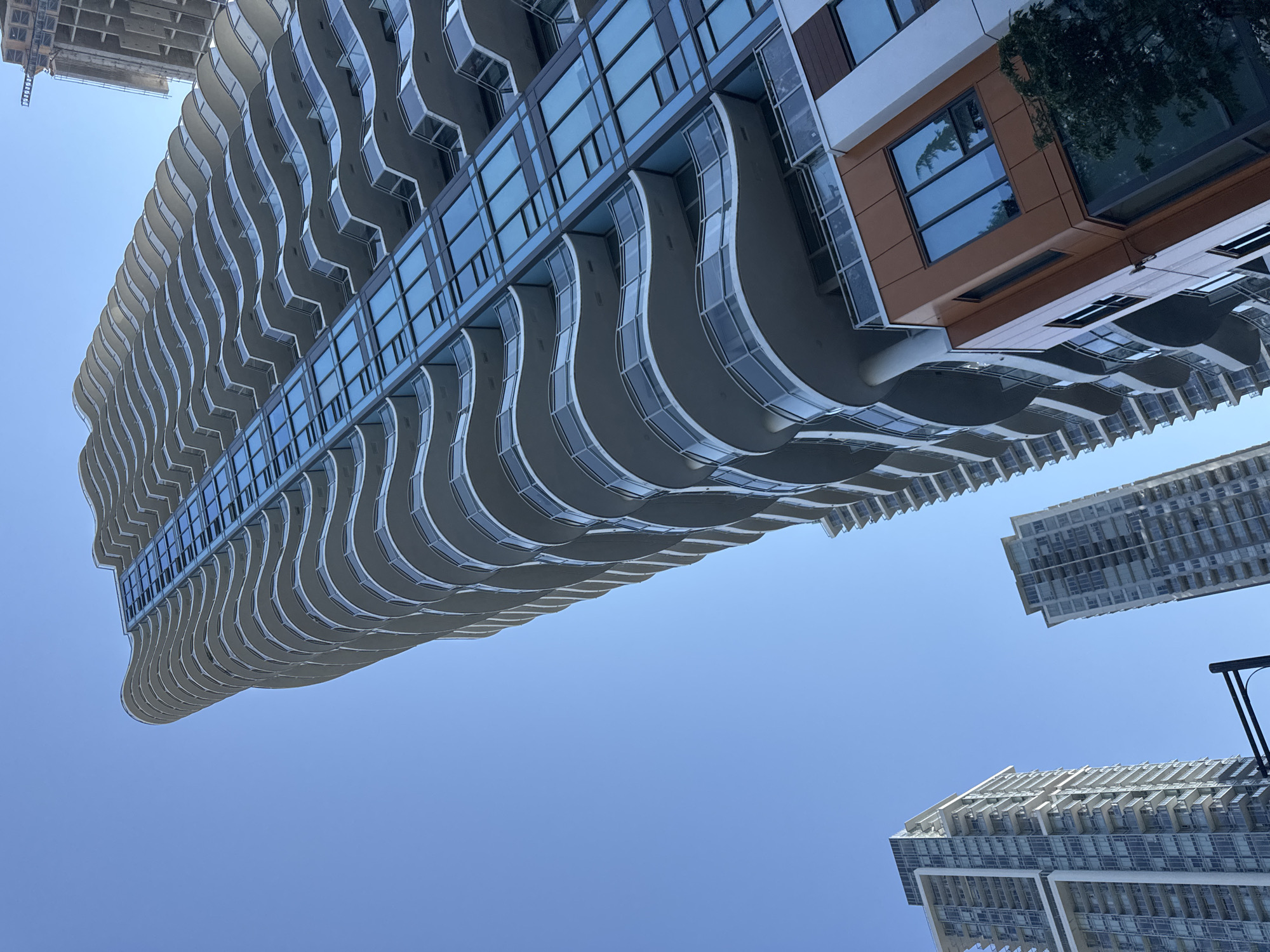 The image shows the exterior of modern high-rise residential buildings in Surrey, Canada, under a clear blue sky. The main subject is a tall, curved skyscraper with numerous balconies that have a wave-like design. The building features a facade of glass windows and white exterior elements. To the left and right, other high-rise buildings are visible, including one that appears to be under construction. The perspective is looking upwards from the ground, emphasizing the scale and height of the structures. The scene is devoid of people or any overt activity.