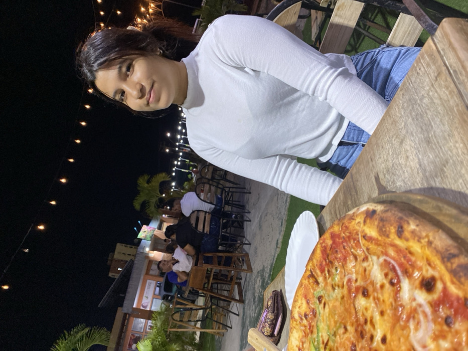 A young woman in a white long-sleeved top sits at a wooden table. A pizza on a wooden board and a plate are on the table in front of her. Behind her, people sit at tables on a grassy patio lit by string lights. A building is visible in the background. The woman looks towards the camera with a slight smile.