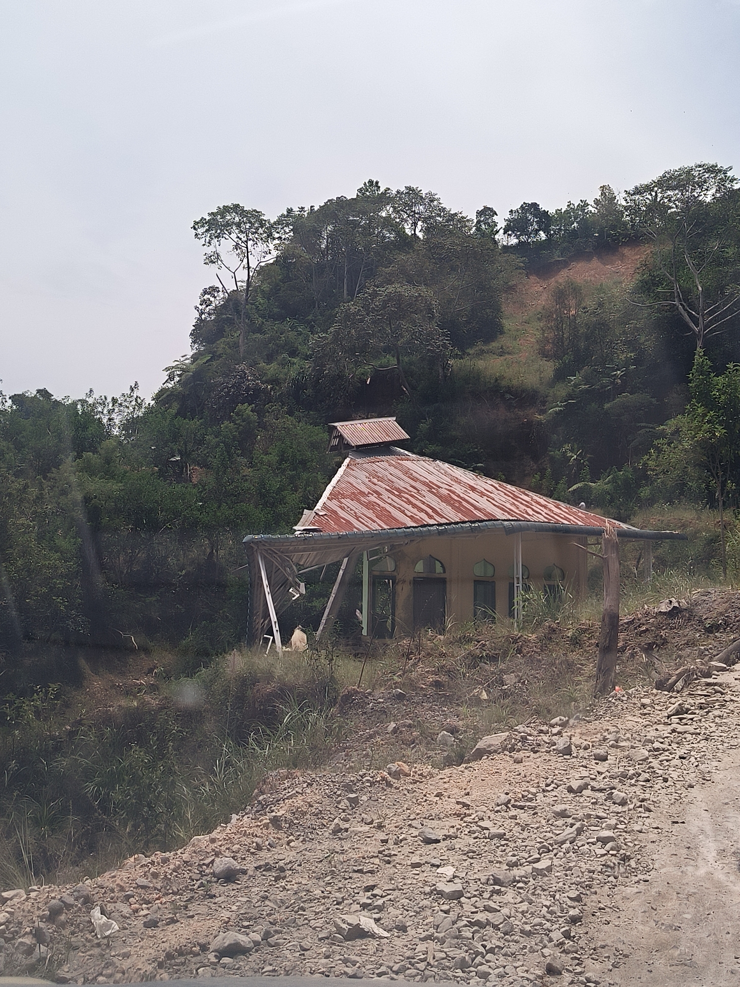 The image shows an abandoned building with a corrugated metal roof in a hilly, forested area. The building is yellow and appears to be in disrepair, with some of the roof supports damaged. There are several windows and doors visible. The surrounding environment is characterized by dense vegetation, including trees and tall grass. The ground in the foreground is covered with gravel and dirt. The sky is overcast. The location is identified as Bintangpermata, Indonesia.