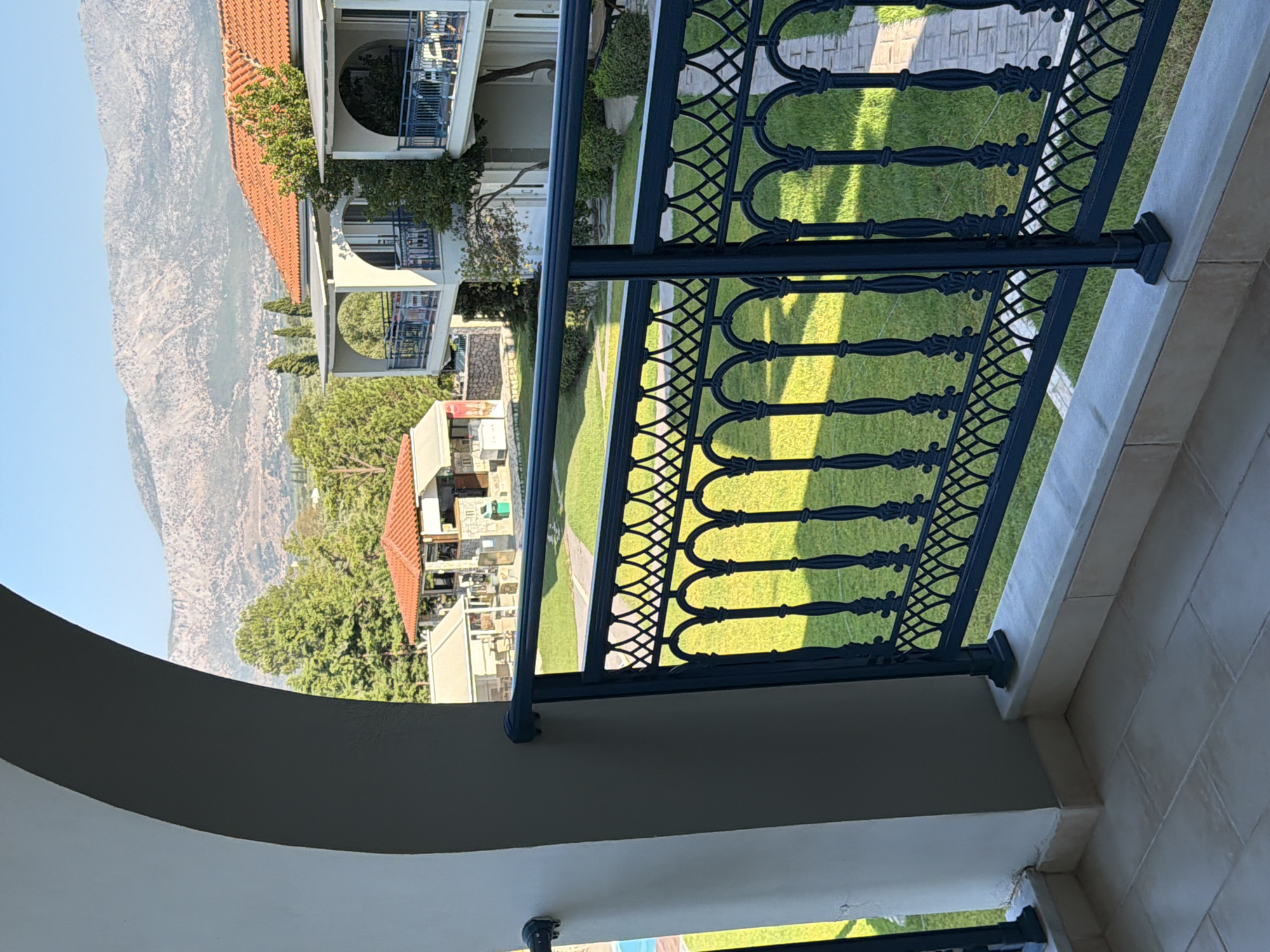 The image shows a view from a balcony looking out over a green courtyard. In the foreground, a dark metal railing with an ornate design runs horizontally across the bottom of the frame. Beyond the railing is a well-maintained lawn with a paved pathway winding through it. To the right, two white multi-story buildings with red tile roofs are visible, featuring balconies with dark railings. In the background, lush green trees and distant mountains under a clear blue sky complete the scene. The location is identified as the Municipal Unit of Alyzia, Greece, suggesting a resort or residential area with a scenic natural backdrop. There are no people visible in the image.