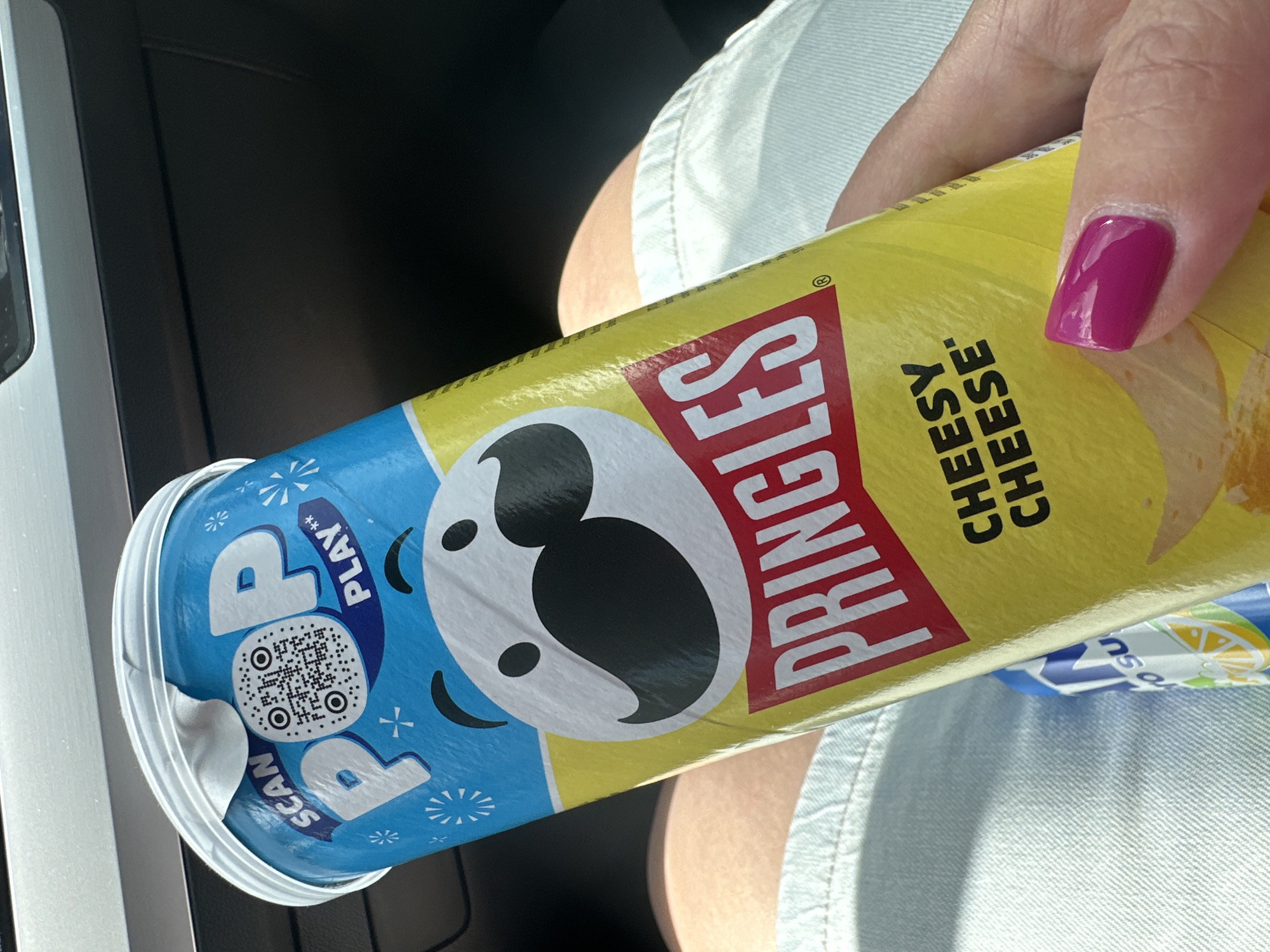 A person is holding a can of Pringles with a cheesy cheese flavor. The can is yellow and has the Pringles logo and mascot on it. The lid of the can is blue and has the words "POP PLAY SCAN" and a QR code on it. The person's hand is visible, and they have bright pink nail polish. The background is out of focus, but it appears to be an indoor setting, possibly a car, given the dark material and lighting. The lighting suggests it's daytime. Another object, a blue can with a lemon slice and what appears to be "SU" is partially visible in the foreground.