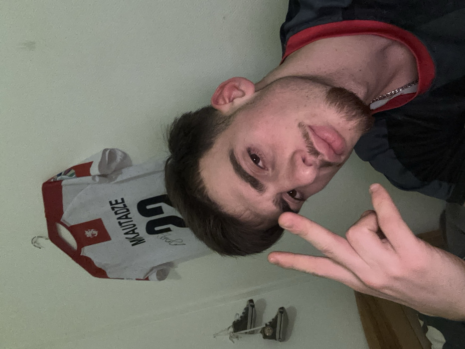 A young man is captured in a selfie-style image, tilted slightly. He displays a hand gesture, resembling a "rock on" or "devil horns" sign. He has short dark hair, a light beard, and appears to be wearing a dark shirt with red trim. In the background, a white sports jersey with red accents is hanging on a hanger. The jersey has the name "MIKAUTADZE" and the number "22" printed on it. Another object hangs below, resembling shoes. The background appears to be a plain wall.