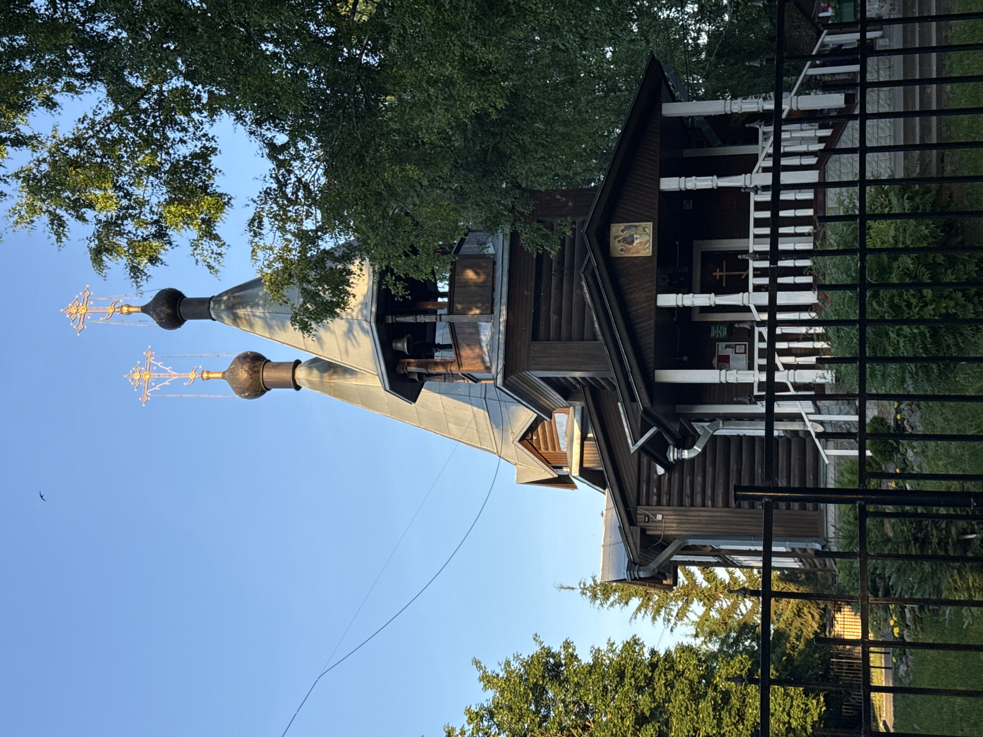 This is a ground-level, eye-level, medium shot of a wooden Orthodox church with two onion domes, located in Vologda, Russia. The church is made of dark logs, with a light-colored porch featuring white pillars and a railing. Above the entrance to the porch, there is a religious icon. Two crosses adorn the tops of the domes, with intricate golden embellishments. A bell tower is visible between the domes. The church is framed by lush green trees on the right and left sides of the image, and a black metal fence is in the foreground, separating the viewer from the church grounds. The sky is a clear, vibrant blue. There are no people or animals visible in the frame. The lighting suggests it is either early morning or late afternoon, with the sun casting warm light on the church and trees.