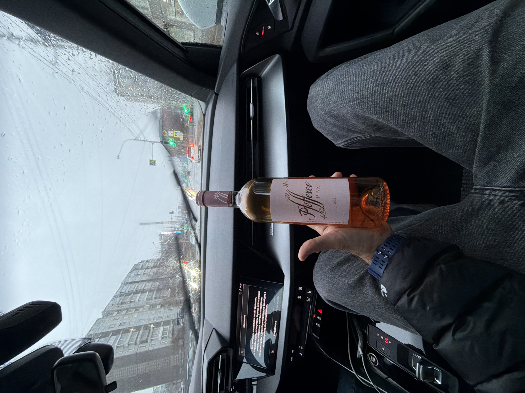 The image shows the interior of a car, focusing on the driver's side. A person is holding a bottle of "Pfeffer PINK 2021" rose wine. The person is wearing jeans, a jacket, and a watch. The car's infotainment screen is displaying information in Russian, with the time "19:30" visible. The windshield shows a rainy urban scene with buildings and traffic lights. Based on the architectural style of the buildings visible through the windshield, the location is likely Krasnoyarsk, Russia.