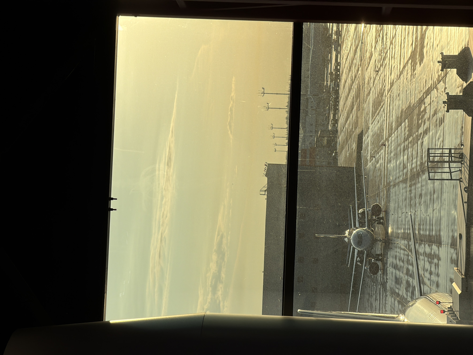 A view through a window at an airport showing an airplane parked at the gate. The sky is overcast and a golden hue suggesting the sun is rising or setting. The tarmac is wet, reflecting the light and nearby structures. The window frame is visible, with a dark interior on one side. Another airplane can be seen in the distance, as well as airport infrastructure such as poles and the terminal.