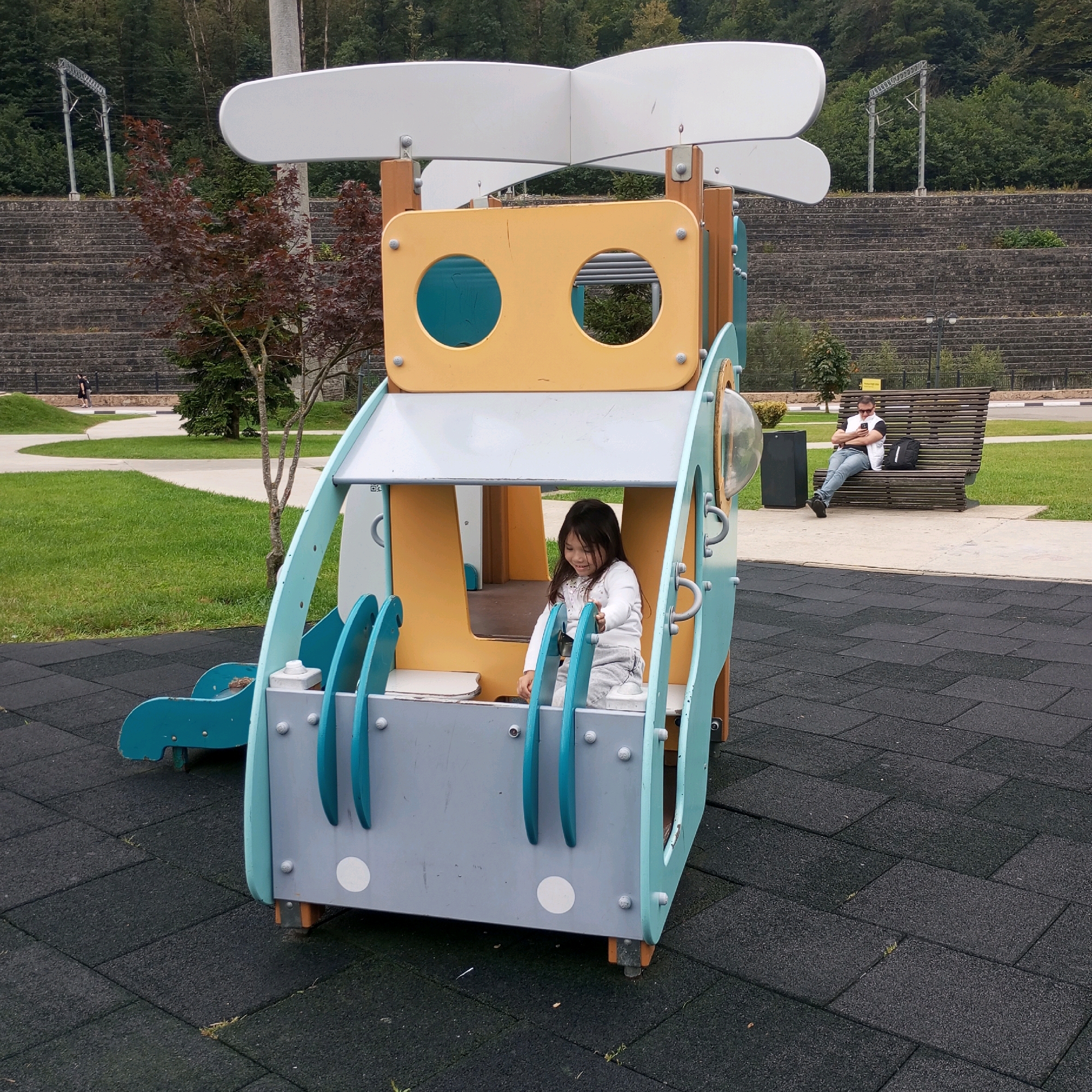 In Estosadok, Russia, a young East Asian girl with dark hair, wearing a white long-sleeved shirt and grey sweatpants, is seated inside a colorful playground structure. The structure has a predominantly yellow and light blue exterior with grey accents and appears to be designed to resemble a vehicle. She is smiling and looking slightly downwards. The playground is situated on a black rubber mat surface, characteristic of children's play areas. In the background, a man wearing a white vest over a black t-shirt, blue jeans, and sunglasses is seated on a slatted wooden bench, looking at his phone. Next to him on the bench is a black backpack. Beyond the bench, a paved walkway curves through a grassy area, and further back, a stone-tiered hillside is visible, topped by dense green trees. Overhead, power lines with tall metal supports stretch across the landscape. The overall scene depicts a public park or recreational area.