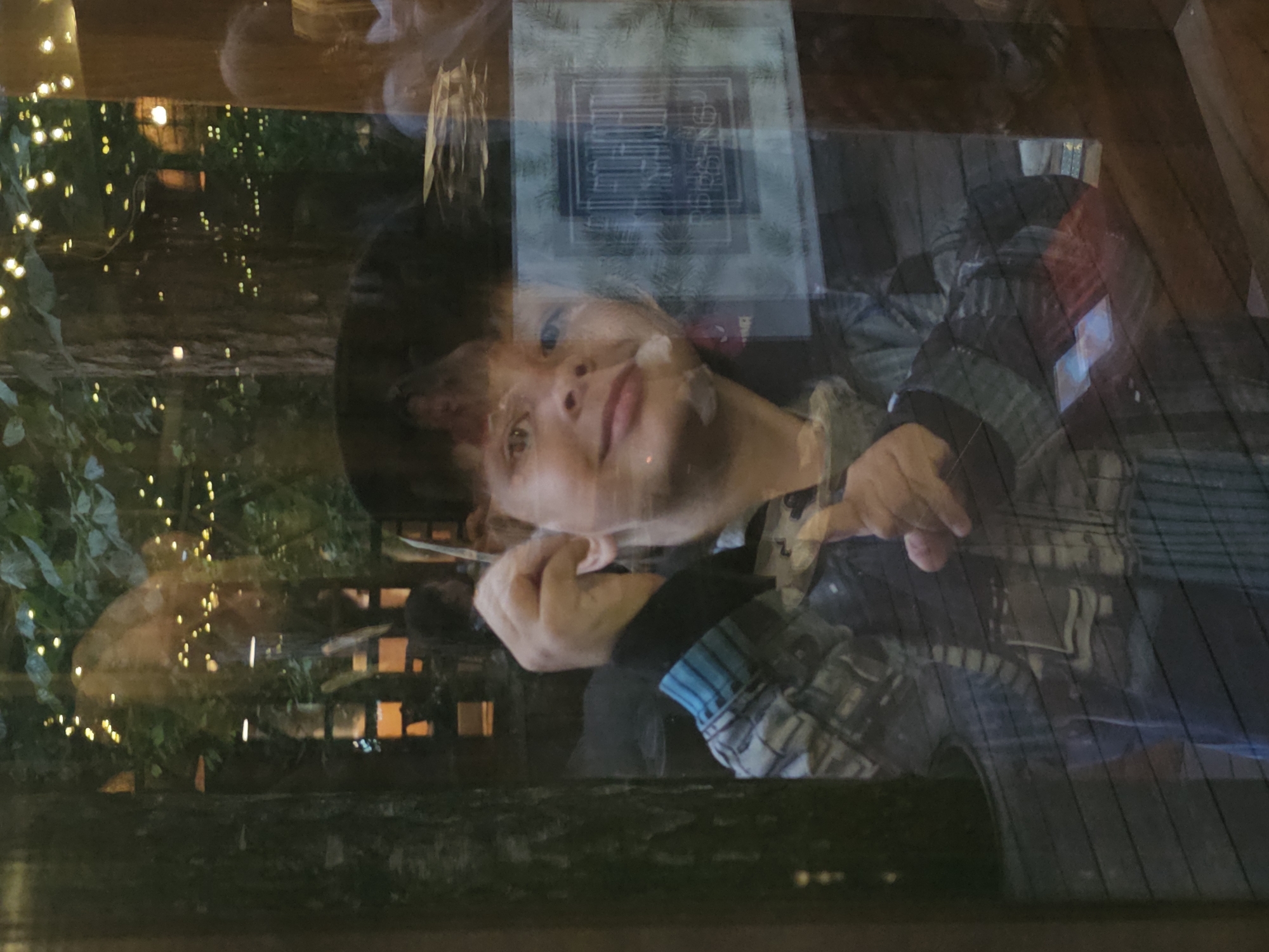 A reflection in a glass window shows a young boy with dark hair, wearing a plaid jacket. He is looking towards the viewer and pointing with his right hand. His left hand is near his ear. In the background, through the glass, trees with fairy lights are visible, suggesting an outdoor setting at dusk or night. The reflection also shows other people and interior elements of a building. The location is specified as Nemchinovo, Russia.