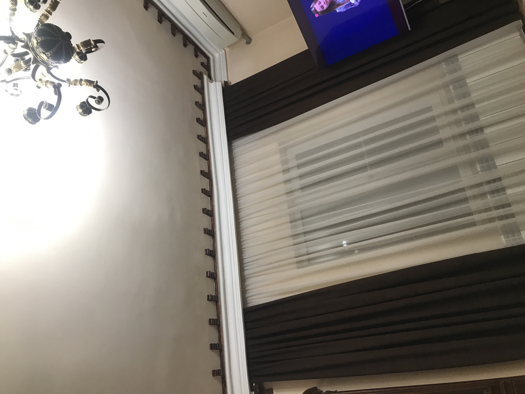 The image shows a view of an interior with various elements:
* **Lighting**: An ornate chandelier is visible on the left side, likely providing illumination.
* **Wall**: A light-colored wall is seen, extending vertically.
* **Window**: A window with horizontal blinds is present, covered with dark curtains above and below.
* **TV**: A television screen is situated in the top right corner.
* **AC unit**: a white AC unit is visible above the wall, close to the chandelier.
Without any additional information or visual clues about the Yerevan cityscape or landmarks, there's no way to conclusively confirm that the location is indeed in Yerevan, Armenia. The image only provides details about the interior space.