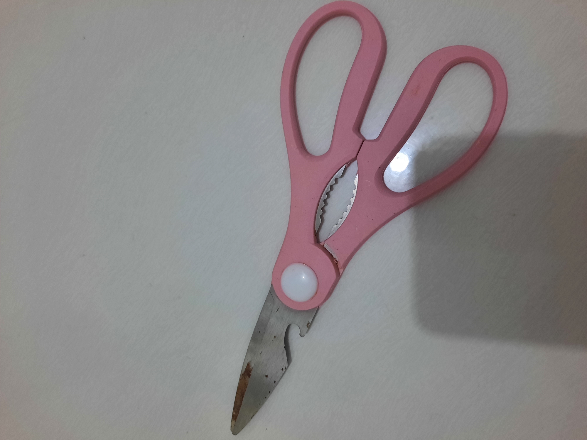 A pair of pink-handled kitchen shears with stainless steel blades rests on a white surface with a subtle texture. The blades exhibit some rust spots. One blade has a serrated edge, and the shears include a bottle opener notch. A white pivot point is visible. The scene is illuminated by overhead light, casting a shadow to the left of the shears. The image appears to be taken indoors in Lhokseumawe, Indonesia, likely in a kitchen or dining area, though no specific landmarks are visible to confirm the exact location within the city.