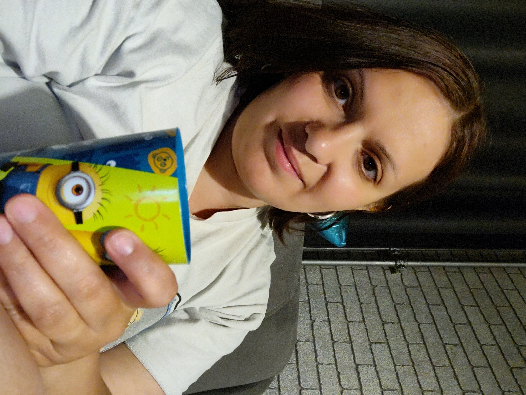 A woman with shoulder-length brown hair holds a cylindrical container decorated with a Minion cartoon character in her left hand. She is wearing a light-colored t-shirt. A blue conical object, possibly a party hat or earring, is visible near her ear. The background consists of a dark surface above and a textured grey surface, resembling paving stones, below. The lighting appears to be artificial, possibly indoor lighting. The image is taken at an angle, as if she is holding the camera herself. There is no contextual information in the image to identify the specific location in Марковский, Russia.