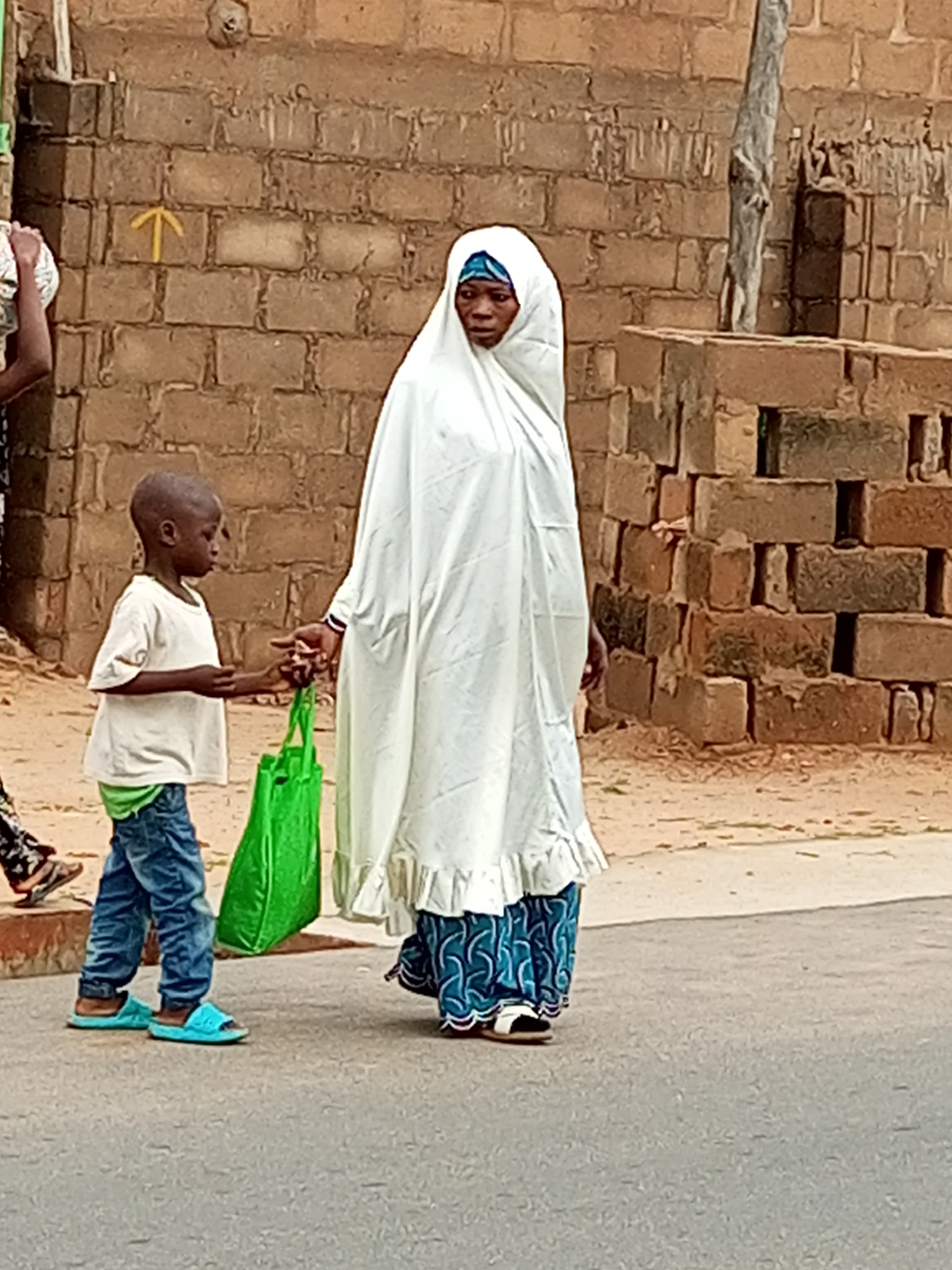 In Tempure, Nigeria, a young boy in a white t-shirt and blue jeans, wearing blue sandals, is walking on a paved road. He is holding a bright green reusable bag in his left hand. A woman, wearing a white headscarf and a blue and white patterned skirt, is walking beside him. They appear to be walking together, with the woman's right hand possibly touching the boy's left hand, or near it. In the background, there is a brick wall constructed from rough, reddish-brown bricks, and a stack of similar bricks. A thin, gray, weathered piece of wood or post stands vertically behind the woman. The ground is a mix of dirt and the paved road. A yellow arrow is painted on the brick wall, pointing upwards.
