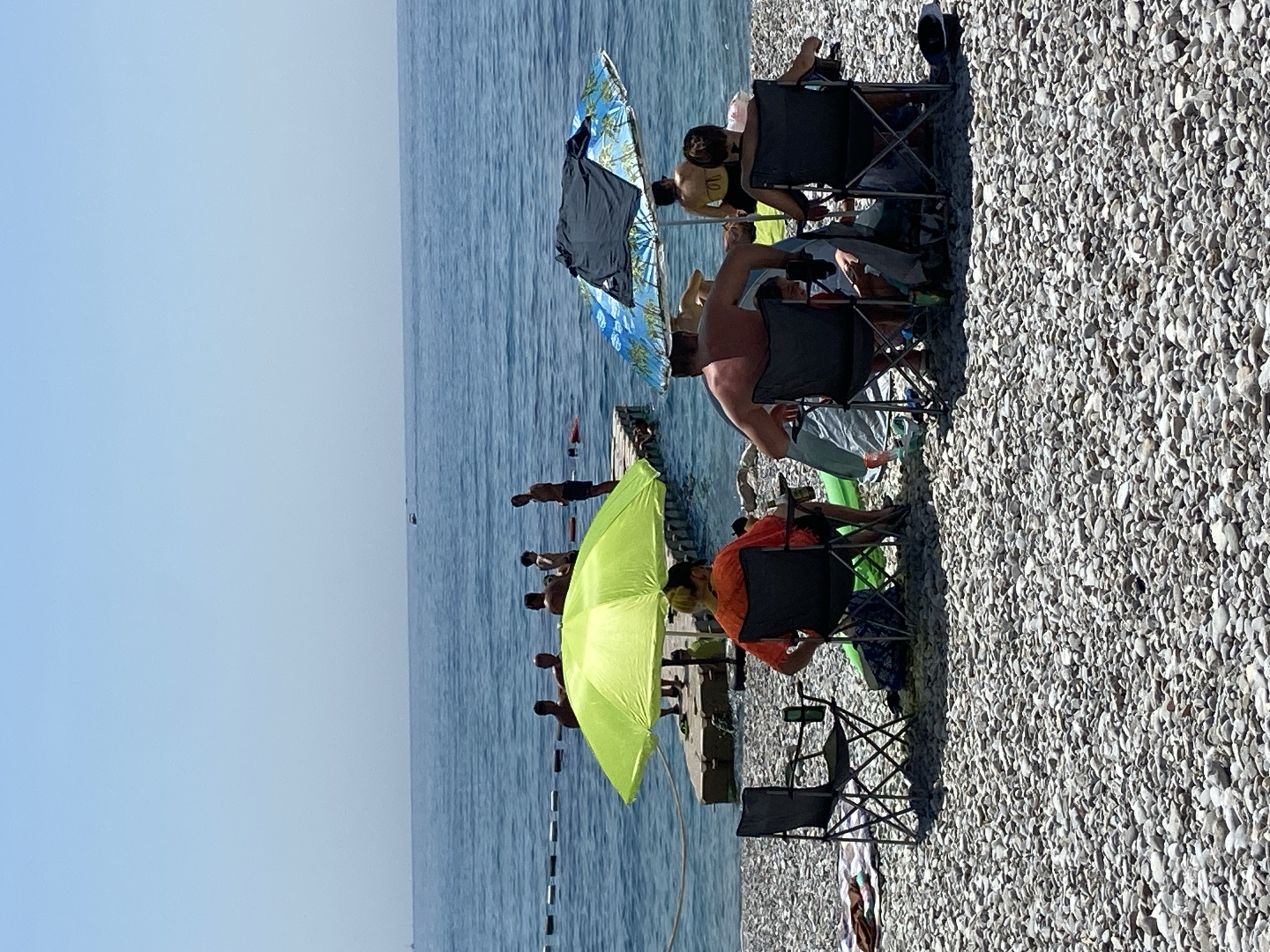The image depicts a sunny day at a pebbled beach in Prijevor, Montenegro. In the foreground, several people are seated in folding chairs under beach umbrellas, facing the water. One umbrella is bright lime green, and the other is blue and white with a palm tree pattern. There are at least five individuals visible in the foreground, some appearing to be adults, others possibly children. Further in the background, several people are standing on a wooden pier and in the shallow water. The sea is calm and a clear blue, with a rope barrier visible in the water. In the far distance, the coastline and a few distant boats can be seen under a clear sky. The overall scene conveys a relaxed atmosphere of a beach day.