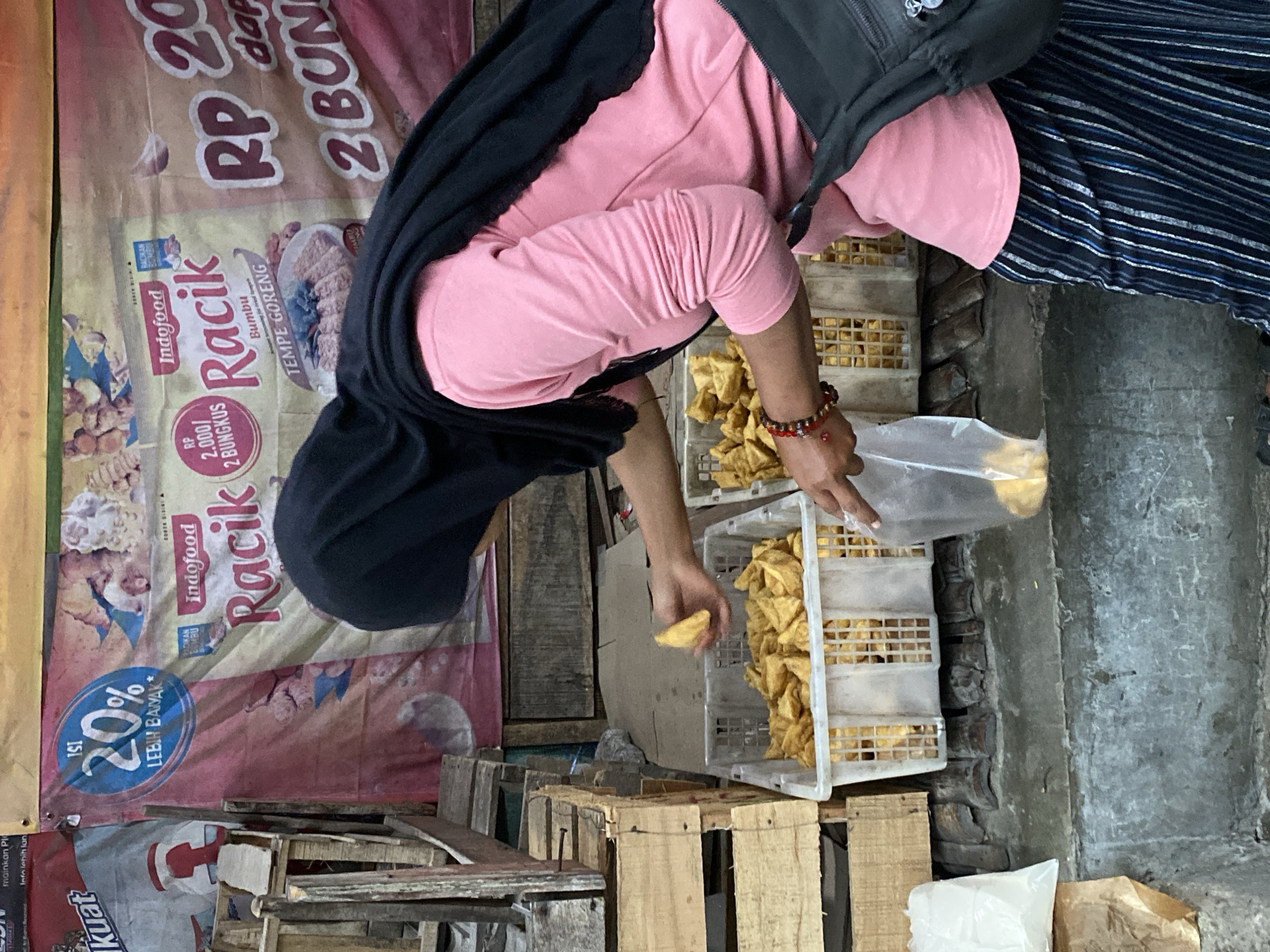 In Ambarketawang, Indonesia, a woman wearing a pink shirt, black hijab, and striped pants is seen at a market stall. She is holding a plastic bag and appears to be selecting a piece of fried food from a large white crate filled with similar items. Another crate of fried food is visible to her right. Behind the crates, a banner displays advertisements for "Indofood Racik" seasoning, featuring promotional text and images. Wooden crates and rustic market structures form the backdrop.