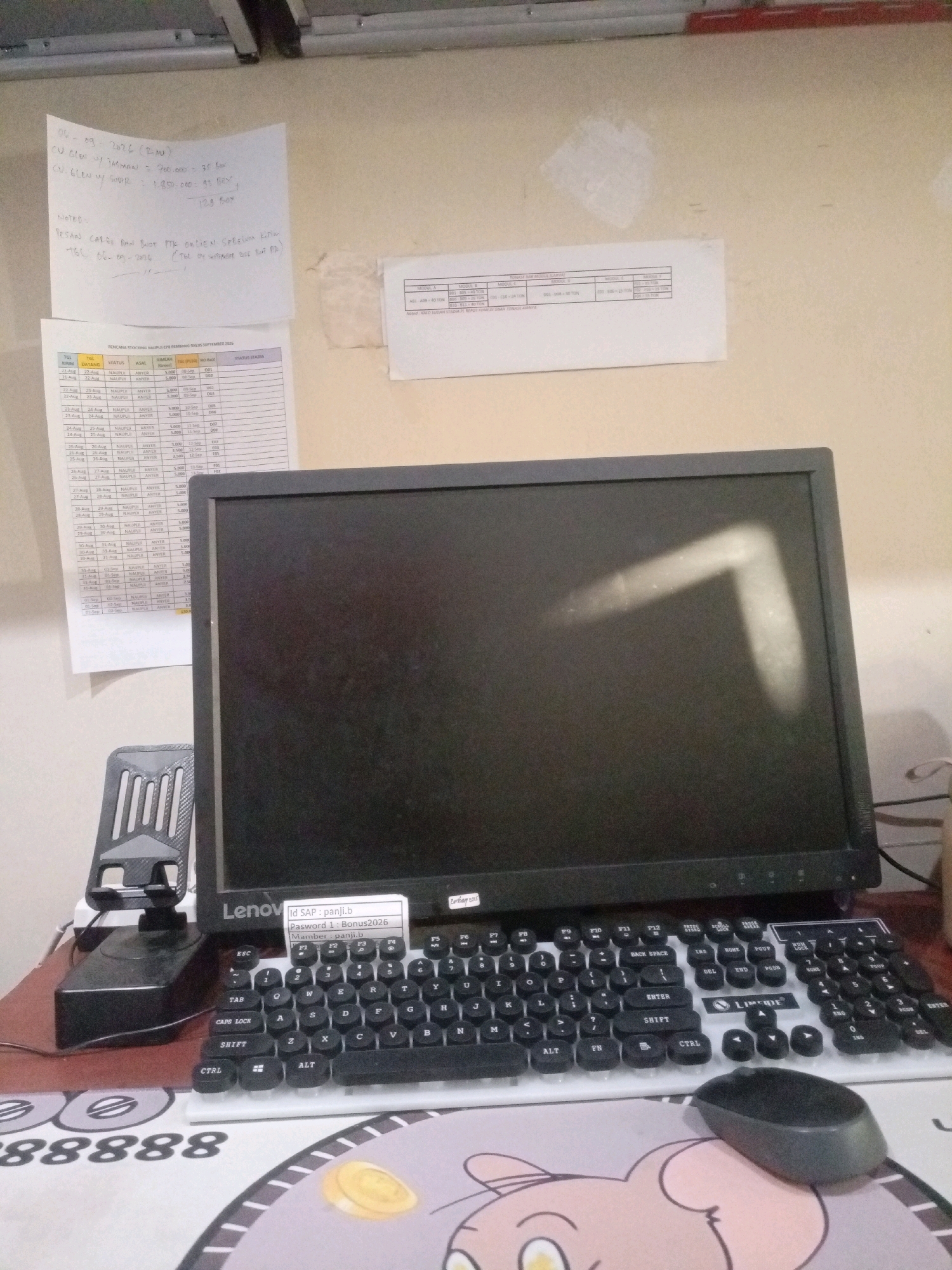 The image depicts a cluttered desk area, likely in an office or home workspace, in the city of Sumbersari, Indonesia. In the foreground, a black computer monitor is the dominant object, positioned centrally. Below the monitor, a white keyboard with black keys is visible, along with a black computer mouse placed on a mousepad featuring a cartoonish design. To the left of the monitor, a black phone stand holds a smartphone.
Several pieces of paper are scattered on the desk. One prominent paper, located to the left and slightly above the monitor, contains handwritten notes in what appears to be Indonesian, detailing some kind of order or request with dates and quantities. Another paper, below the first, is a printed table with columns that include "TGL", "DATANG", "STATUS", "ASAL", "JUMLAH", "TBN (PULAU)", and "STATUS STADIA". This table seems to be a logistics or delivery manifest. A third, smaller paper is attached to the monitor's bezel, displaying login credentials.
The background shows a plain, off-white wall with some smudges and scuff marks, suggesting a lived-in environment. The lighting appears to be artificial, casting shadows and highlighting certain areas, particularly a faint, angular streak of light on the monitor screen itself. The overall impression is one of a functional, albeit somewhat disorganized, workspace. There are no people visible in the image.