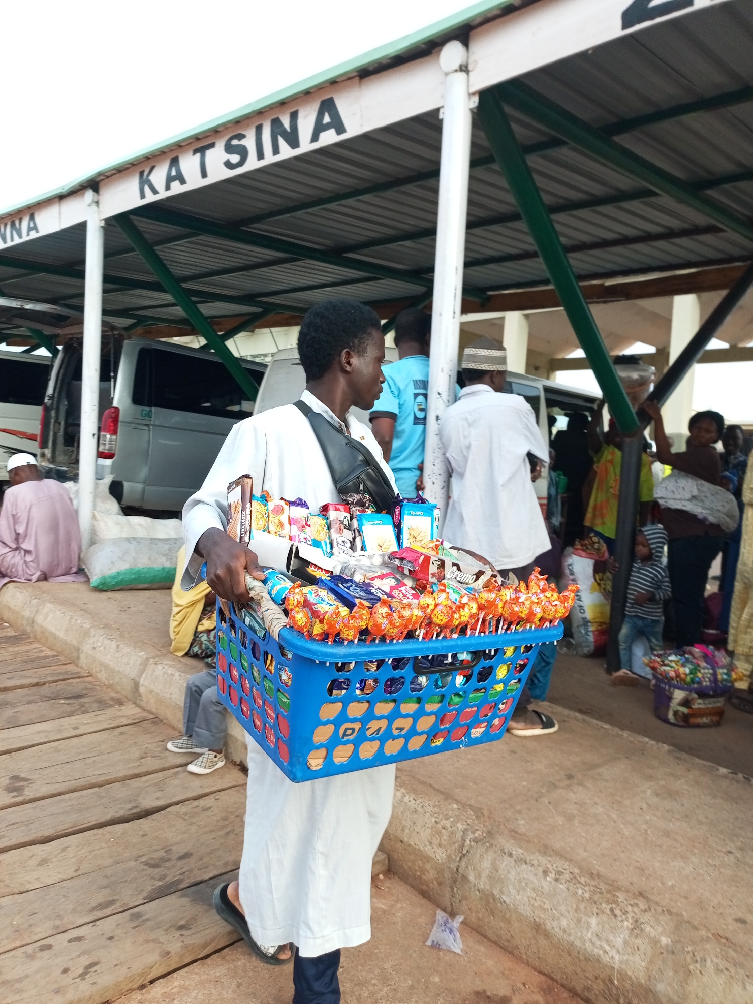 A Black male vendor, wearing a white robe and a black fanny pack, walks toward the viewer while carrying a blue plastic basket overflowing with colorful candy and snacks. He is standing on a wooden platform next to a concrete curb. In the background, several people are visible, including individuals sitting on the curb and standing near vehicles. Above them, a white structure with green trim is visible, featuring the word "KATSINA" in large black letters. The overall scene appears to be an outdoor market or transport hub in Gombe, Nigeria.