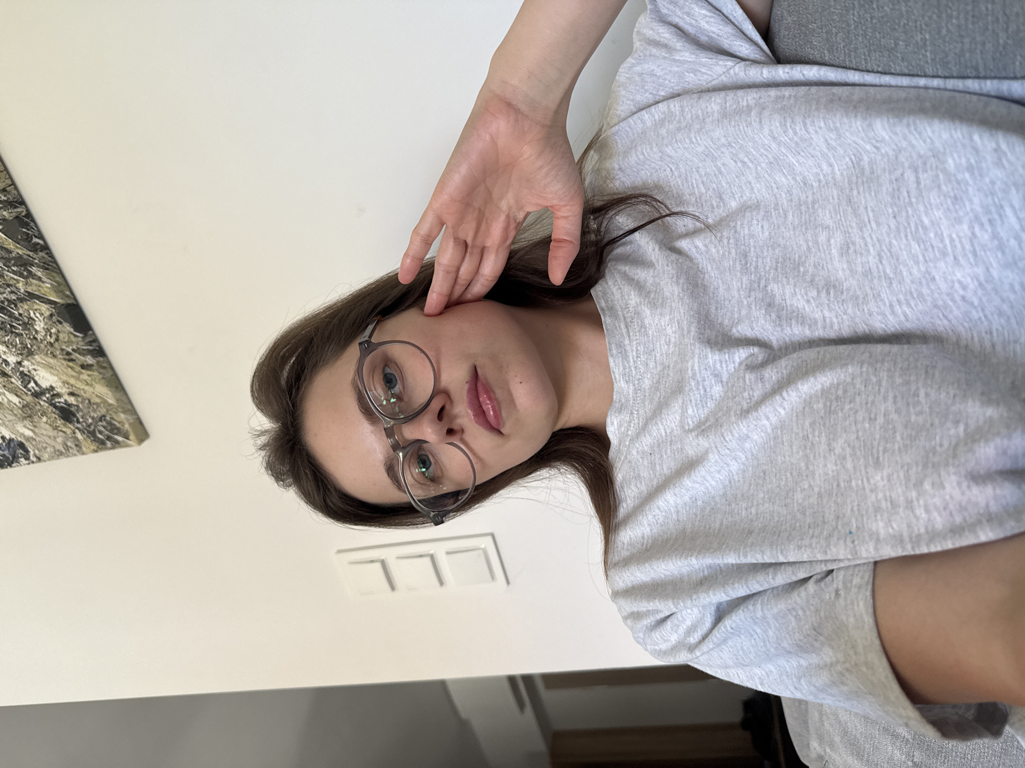 A young woman with long brown hair and glasses is sitting indoors, looking at the camera. She is wearing a light gray t-shirt. Her right hand is raised and touching her face. The background is a plain white wall with a light switch panel. A framed picture with a nature scene is hanging on the wall above her. The image appears to be a selfie taken in a domestic environment. There is no visible indication of the location being in Szklarska Poręba, Poland.