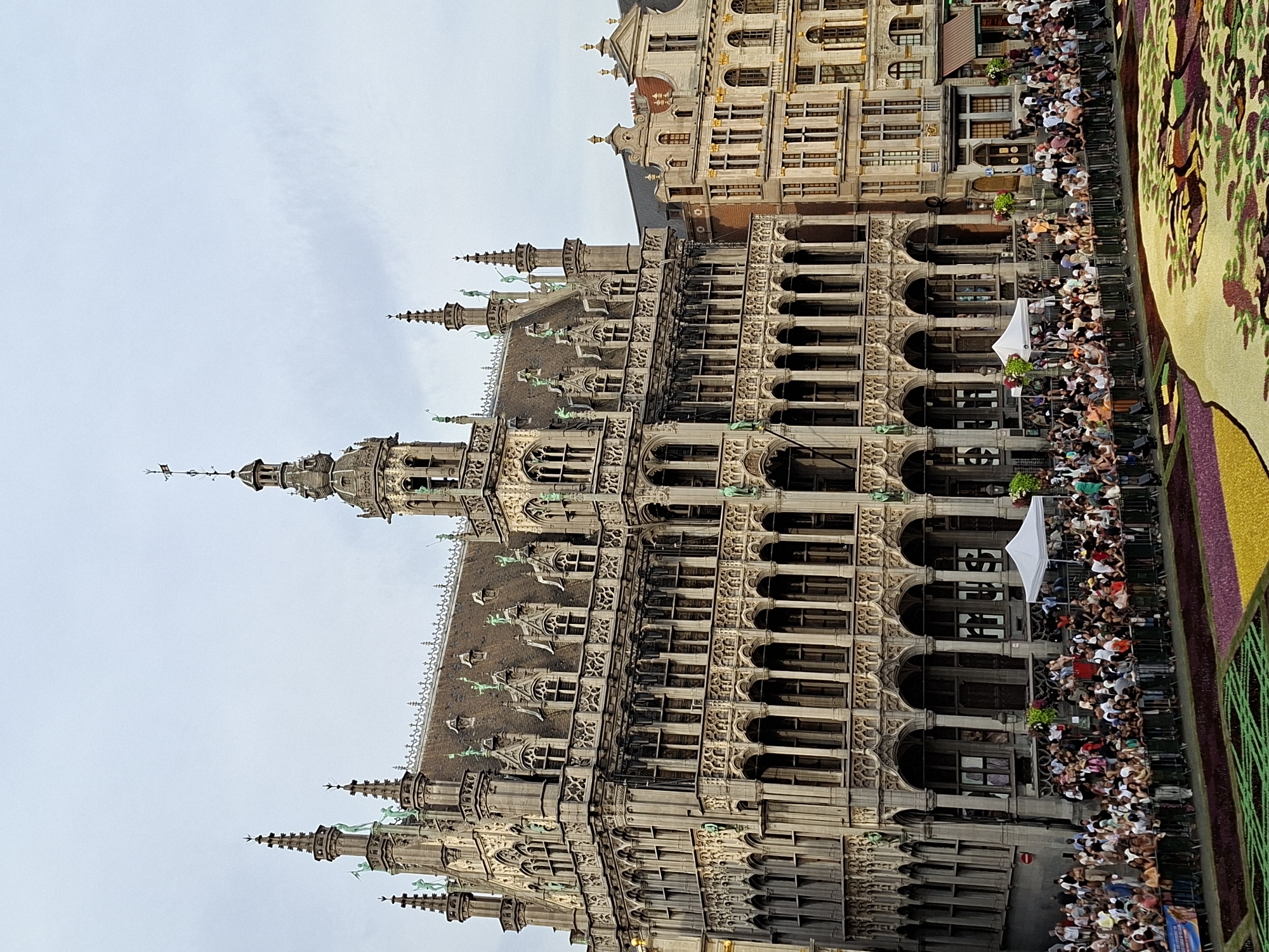 The image depicts the Grand Place in Brussels, Belgium, featuring the ornate facade of the City Hall. A large crowd of people is gathered in front of the building, separated by metal barriers. In the foreground, a vibrant flower carpet with intricate patterns is visible, with sections of purple, yellow, and green. The sky is overcast. The architecture of the City Hall is characterized by its Gothic style, with numerous spires, statues, and arched windows. Adjacent buildings, also displaying historical architectural styles, are visible to the right of the City Hall. White tents are set up near the building, and some individuals appear to be observing the flower carpet from the upper levels of the surrounding structures.