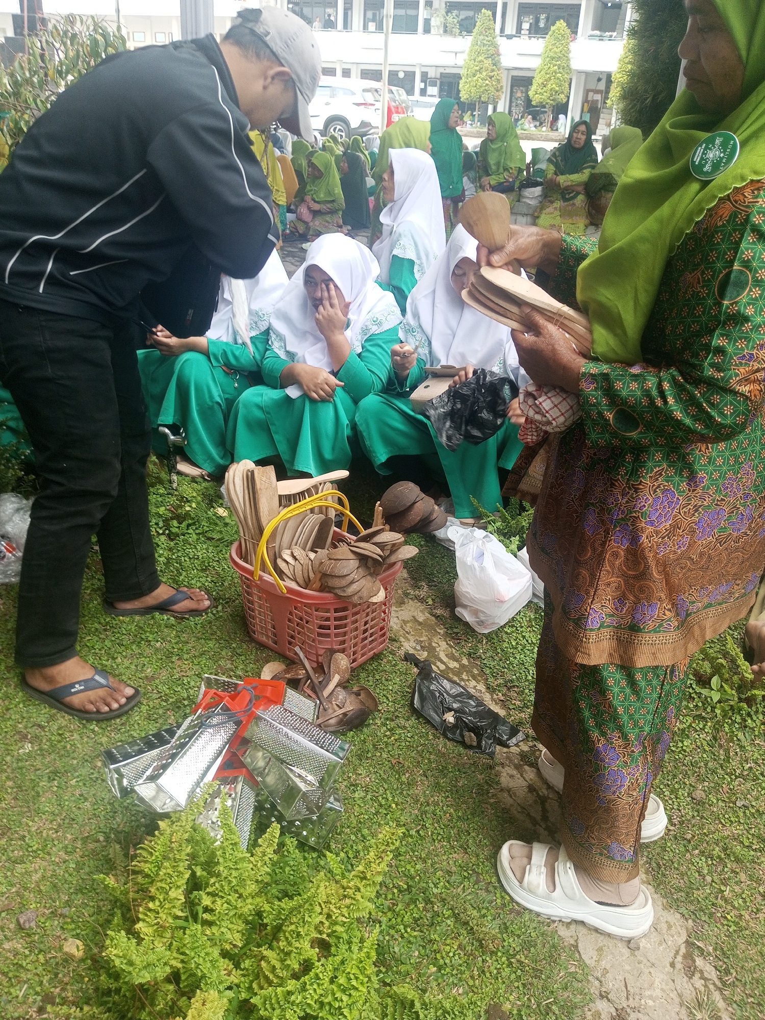 In Klaseman, Indonesia, outdoors on a grassy area with some concrete, a group of people, mostly women wearing green and white hijabs and traditional attire, are gathered. A man in a black jacket and black pants, wearing a grey cap and dark flip-flops, stands to the left, looking down. In the center and right, women are seated or standing. One woman, wearing a green headscarf with a "MUSLIMAT" emblem, holds several wooden utensils, possibly spoons and spatulas. A red basket filled with more wooden utensils is placed on the ground in front of her. To the right of the basket, a white plastic bag and a black plastic bag are visible. In the foreground, to the left, a pile of shiny silver graters is arranged on the grass, partially obscured by a lush green bush. Another woman in white sandals stands on a concrete patch to the right, facing away from the camera. The background shows buildings and trees, suggesting an outdoor public space. The scene appears to be a casual gathering or a market stall.