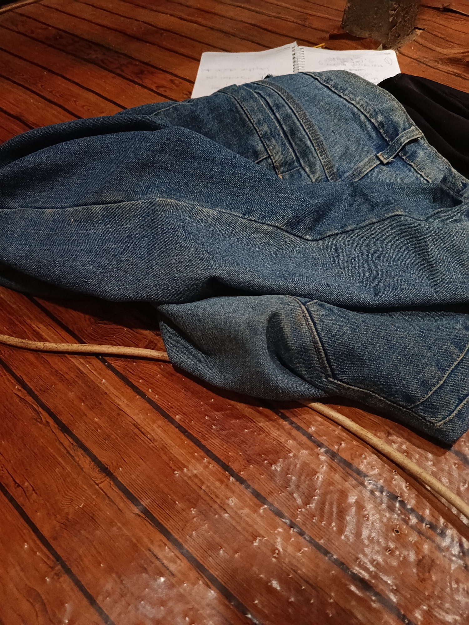 A pair of blue denim jeans is casually draped over a wooden surface that appears to be a table or floor, with visible wood grain and dark lines suggesting planks. A thin, light-colored stick or rod lies across the jeans and the wooden surface. In the background, a portion of a white notepad with writing is visible, along with a dark fabric, possibly clothing. The scene is indoors, with ambient lighting that creates shadows and highlights on the textured surfaces. The specific location in Karangmojo, Indonesia, is not discernible from the visual information provided.