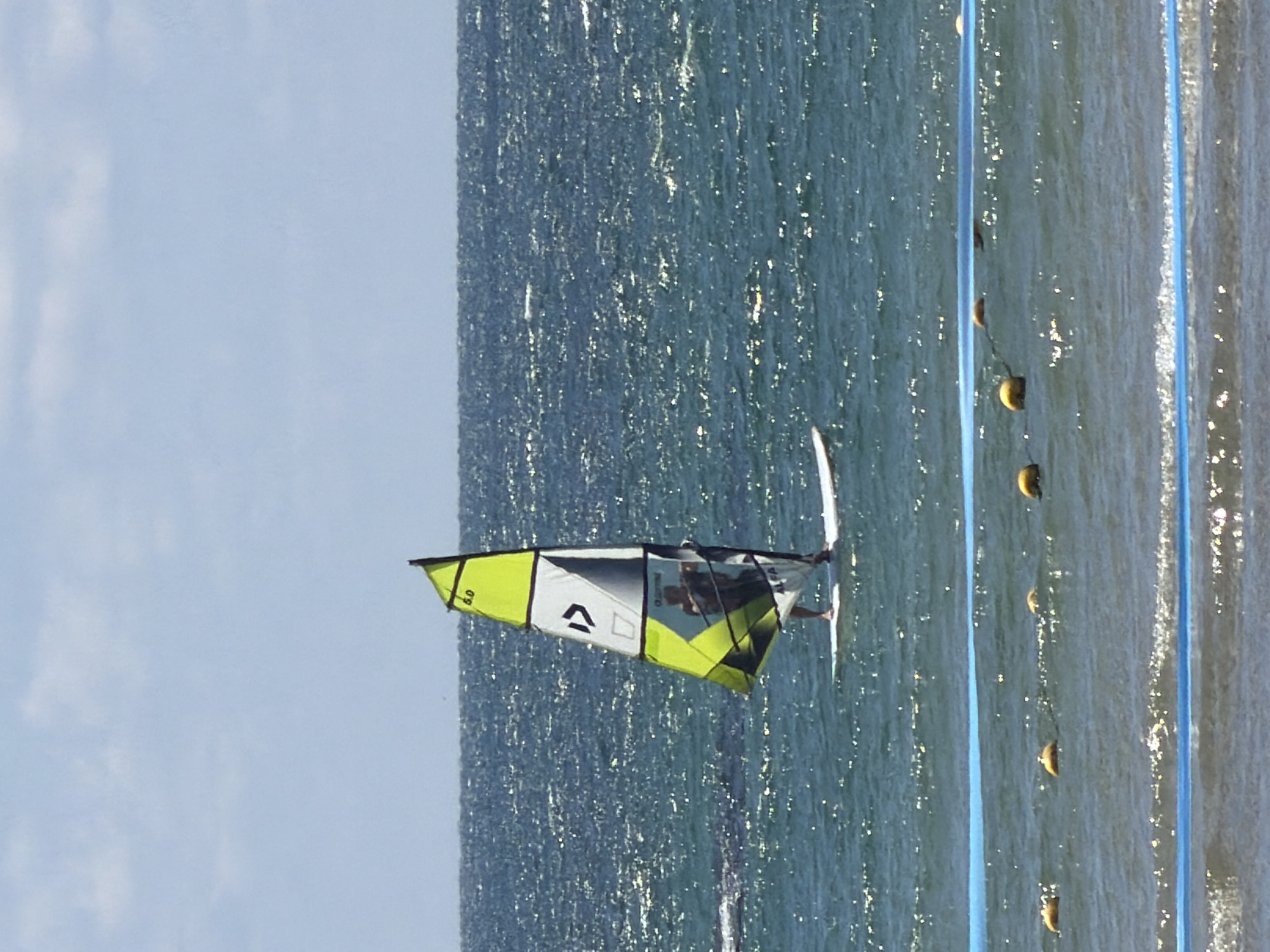 The image depicts a windsurfer on the Mediterranean Sea, likely off the coast of Tel Aviv, Israel. The windsurfer is riding near a lane marker, which consists of yellow floats attached to a blue line, extending along the water. The sail is predominantly green and white with black accents. The sky is visible above the water, showing partial cloud cover. The sea appears choppy with glints of sunlight reflecting off the surface.