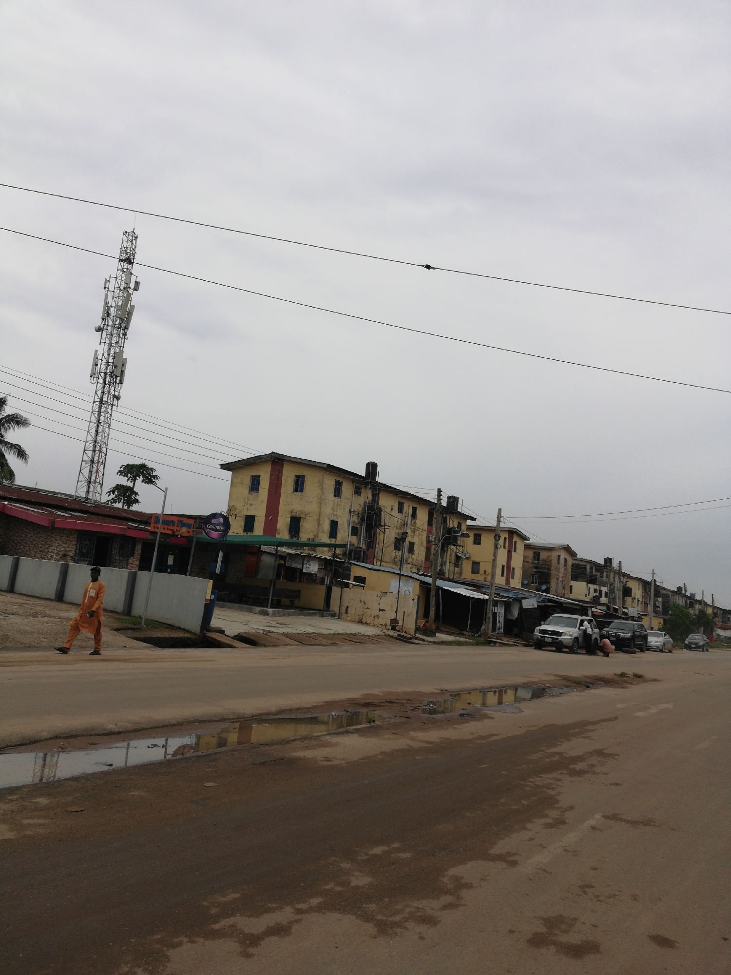 The image shows a street scene in an urban environment. A tall telecommunications tower is visible on the left. The buildings lining the street are multi-story and appear somewhat weathered. Several cars are parked along the side of the road. A man in a long-sleeved orange outfit is walking along the side of the street. Puddles of water are visible on the surface of the street. The sky is overcast.