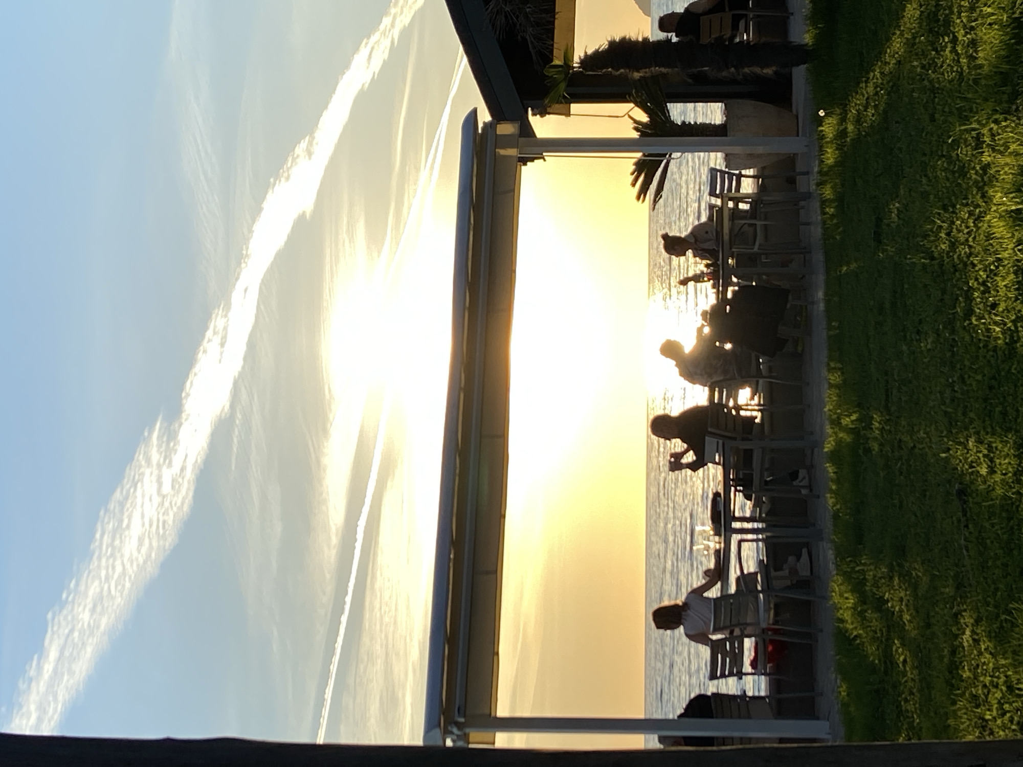An outdoor bar or restaurant by the sea in Bar, Montenegro, is captured at sunset. The sun, low on the horizon, casts an intense golden light across the water, silhouetting the patrons and parts of the establishment. Several people are seated at tables and chairs, facing the water, their figures appearing as dark outlines against the bright backdrop. Some tables have glassware, suggesting they are enjoying drinks or a meal.
The bar structure, featuring a modern design, is visible on the left, with some potted plants or small trees adding to the decor. To the right of the seated area, a patch of vibrant green grass is bathed in the warm light of the setting sun. The sky above is a gradient of bright yellow near the sun, transitioning to a soft blue further away. Distinct white contrails or cirrus clouds streak across the sky, with one prominent, long contrail cutting diagonally across the upper left. The vast expanse of water reflects the sun's intense glow, creating a shimmering surface. The overall atmosphere suggests a serene and relaxing evening gathering. No discernible text is visible.