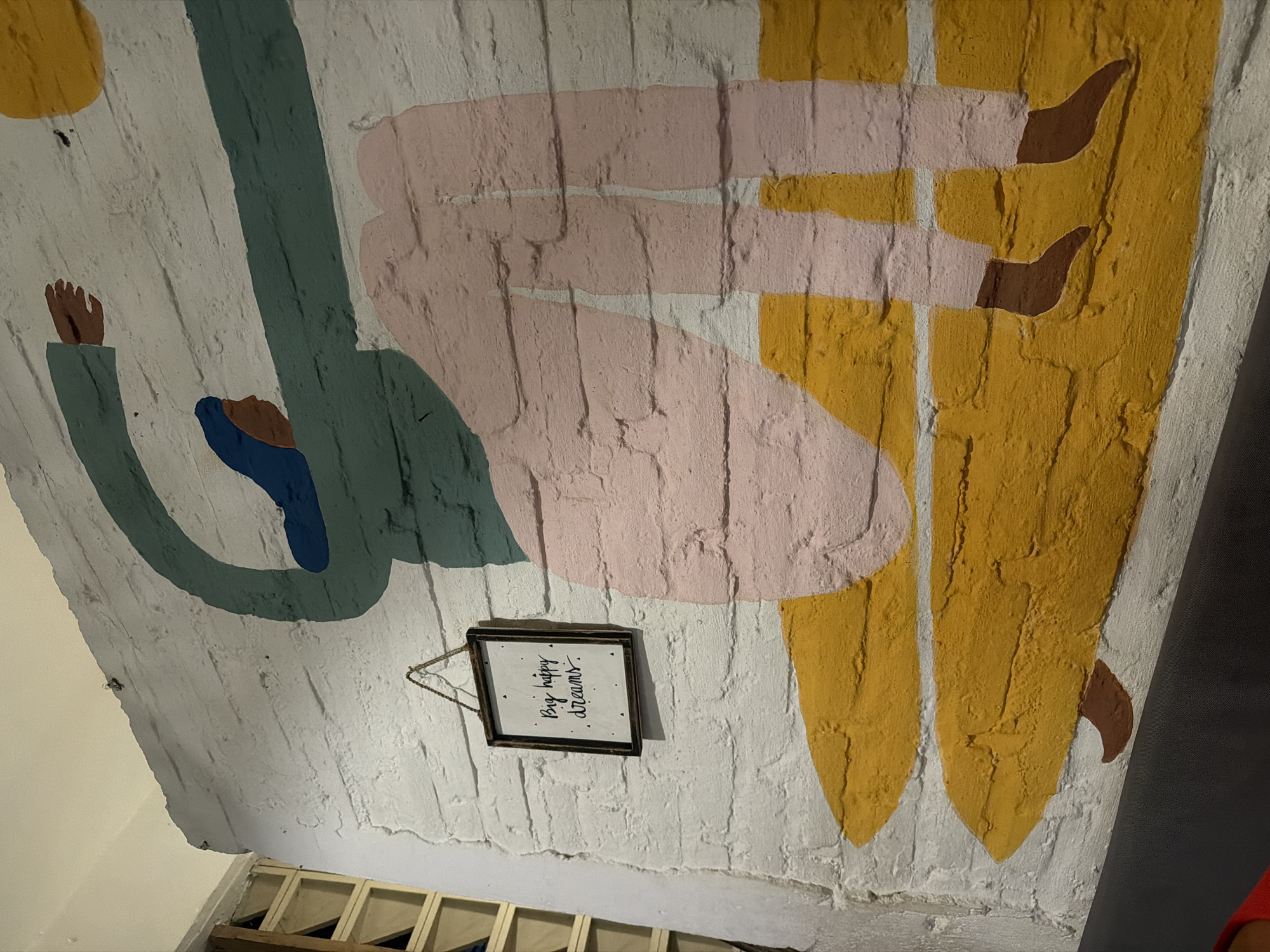 This media file captures an artistic mural painted on a textured white brick wall or ceiling in Kuta, Indonesia, viewed from below looking upwards. The mural features abstract, stylized human figures rendered in flat, block-like colors.
One figure, predominantly in a teal/dark green hue, appears to be reclining or sitting, with a blue accent possibly indicating clothing or another body part, and brown hands. Another figure, sketched in pink, seems to be lying down or stretching across the surface. A third figure, or perhaps parts of another, is depicted in mustard yellow, with brown details suggesting feet or shoes. The overall impression is one of relaxed, almost dream-like postures.
Below the main mural, centered in the image, a small, black-framed sign hangs from a string. The text clearly visible on the sign reads: "Big happy dreams." The backdrop for the mural is the uneven, whitewashed brickwork, which adds depth and texture to the painted forms.
In the lower portion of the image, hints of the surrounding environment are visible: exposed wooden beams or framework underneath the painted surface on the left, a dark grey or black wall/surface on the far right, and a glimpse of a red garment in the bottom right corner. The lighting appears warm and somewhat dim, suggesting an indoor setting like a cafe, restaurant, or a cozy room. The overall atmosphere conveyed by the mural and its text is one of positivity, relaxation, and aspiration.
