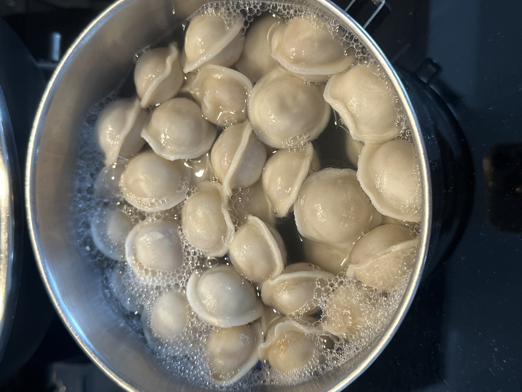 A close-up, overhead shot shows dumplings boiling in a pot of water. The pot is made of brushed metal and sits on a black stovetop. Bubbles surround the dumplings, which appear to be filled with a light-colored ingredient. The dumplings have a characteristic pleated edge. The image is well-lit, with some reflections on the surface of the pot and the water. There is no indication of the location in Vsevolozhsk, Russia.
