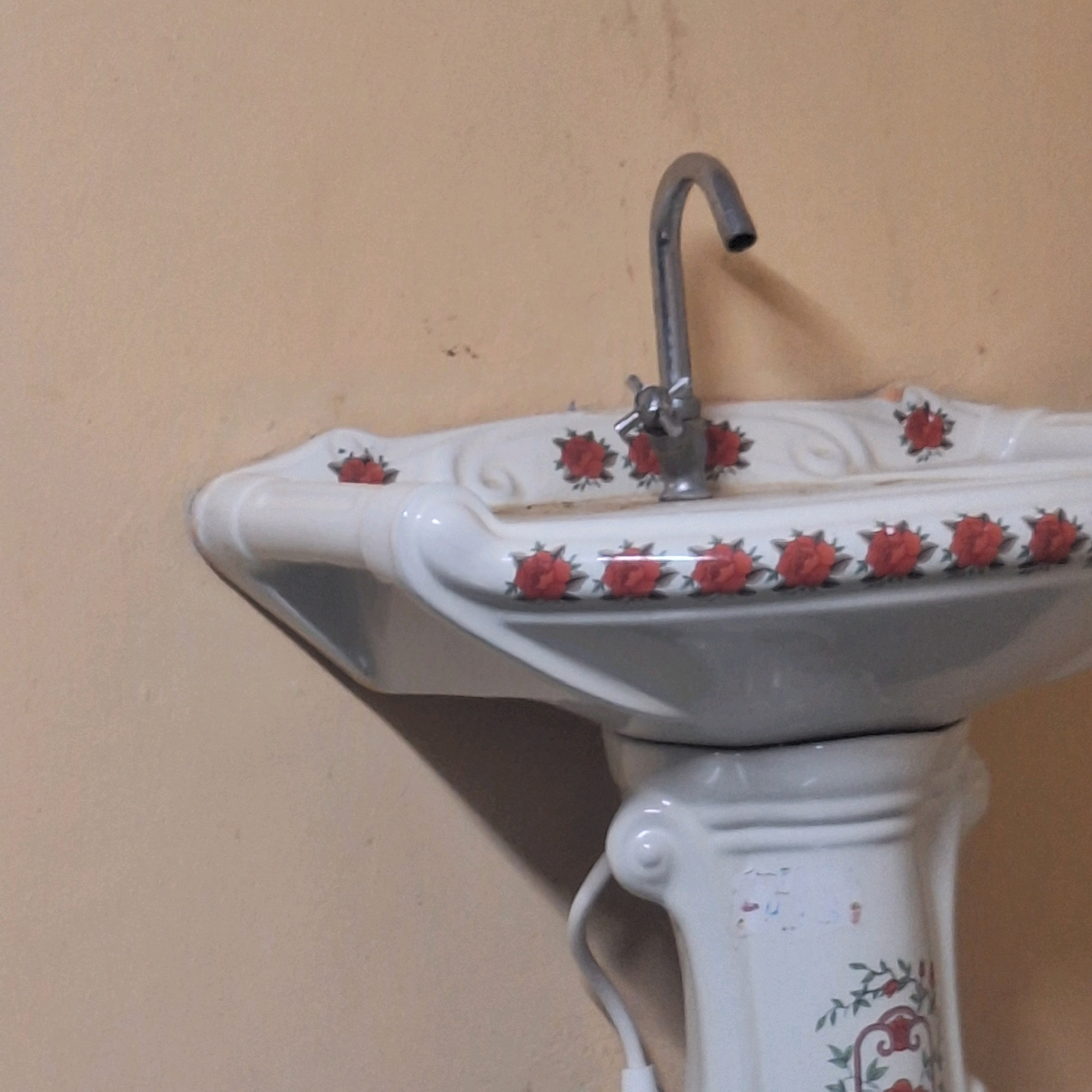 The image shows a close-up of a white pedestal sink with a metallic faucet. The sink has a decorative pattern of red roses and green leaves. The wall behind the sink is a light beige color and appears slightly textured. The lower portion of the sink, the pedestal, is also visible and continues the rose and leaf decoration. There is a white tube or hose extending from the sink's base, partially visible in the lower right of the image. The lighting suggests an indoor environment. There are no people or animals present in the image. The location in Tadasa, India, is not discernible from the visual information provided in the image.