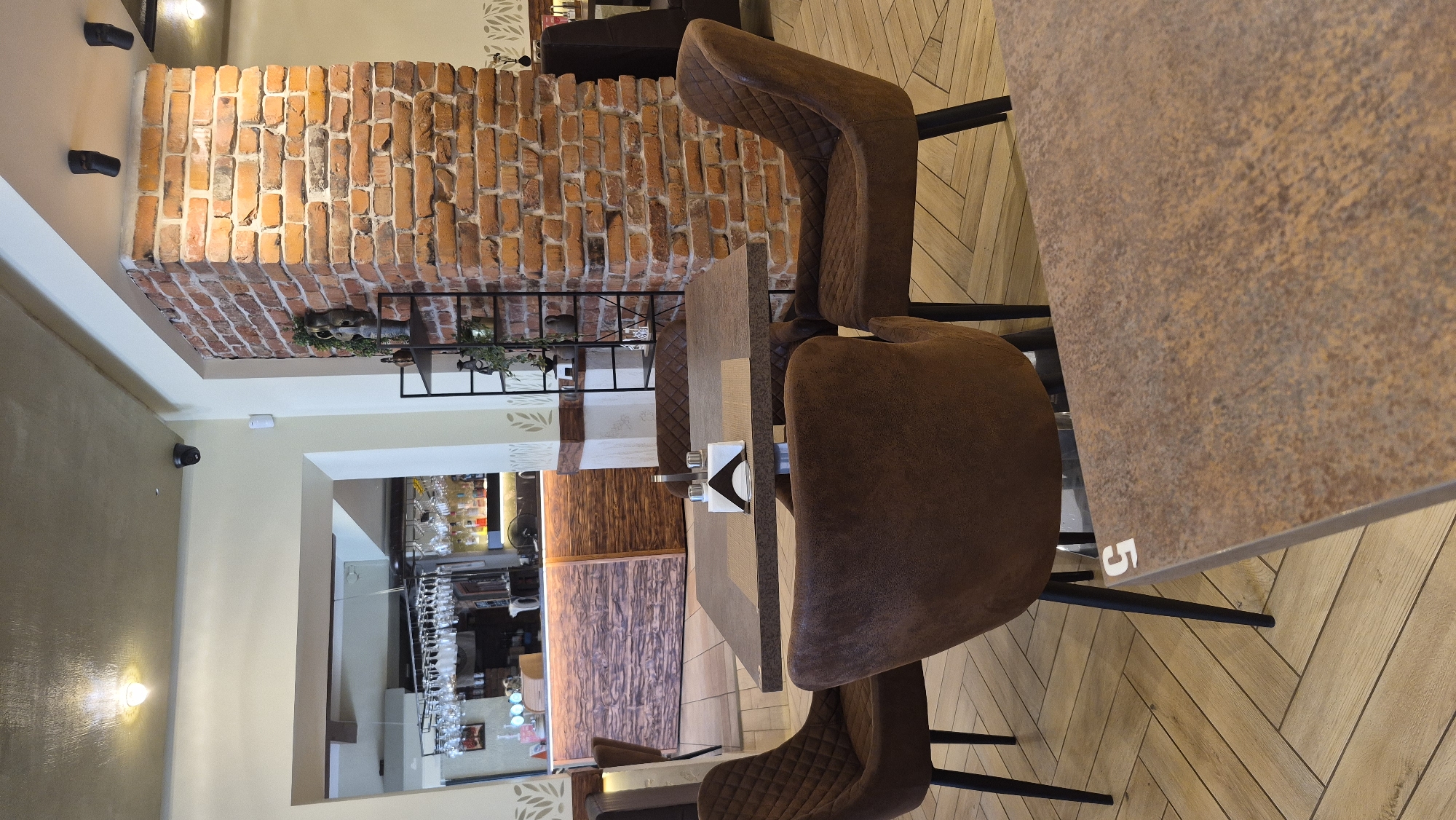 The image shows the interior of a restaurant in Daugavpils, Latvia. In the foreground, there are several brown upholstered chairs arranged around a table. The table has a speckled brown surface and is set with a napkin dispenser and a salt and pepper shaker. In the midground, a brick pillar is visible, and behind it, a bar area with bottles and glasses on shelves can be seen. The floor is tiled with a wood-grain pattern. The overall ambiance suggests a casual dining establishment.