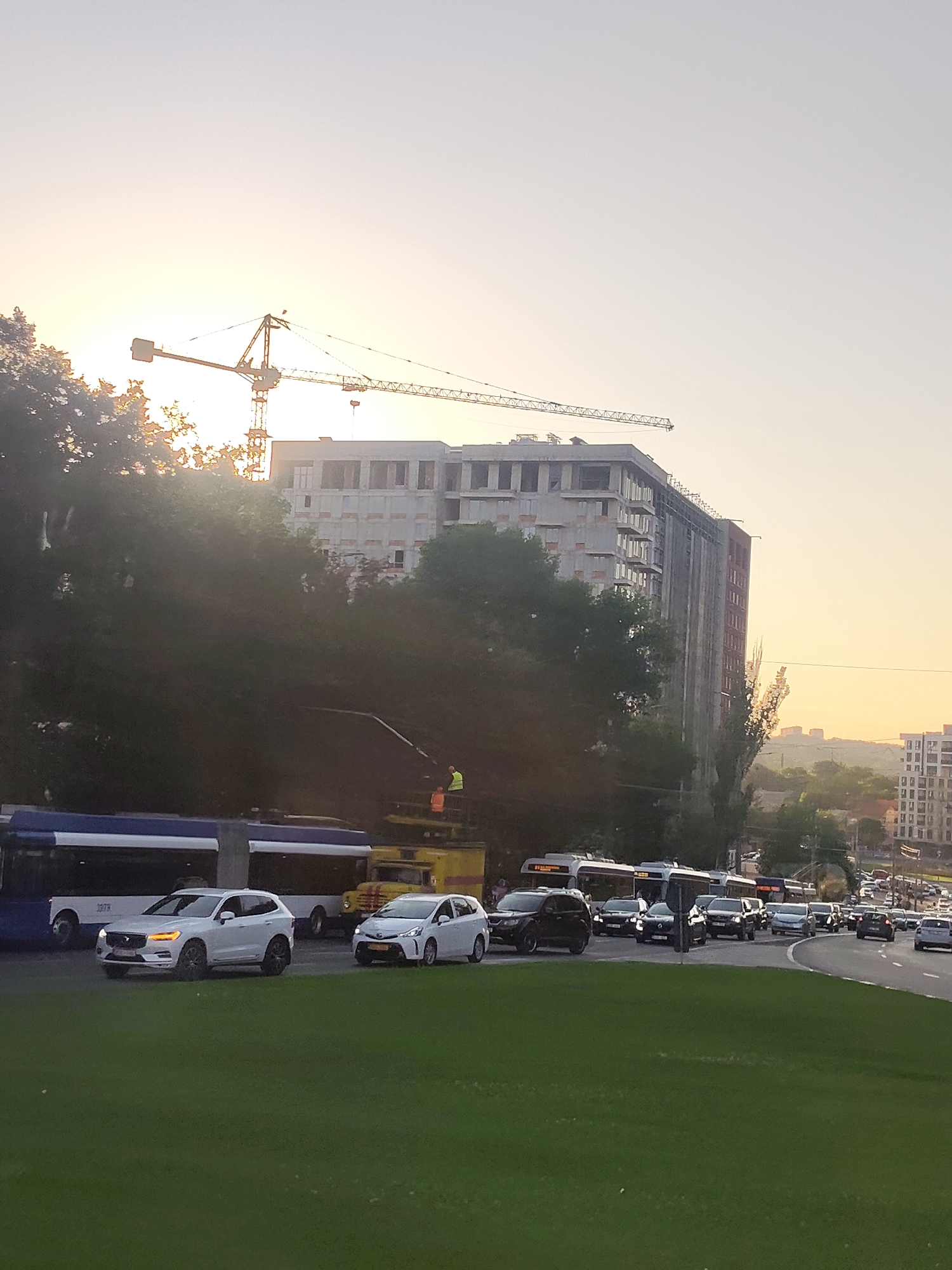 A wide-angle, eye-level shot captures a street scene in Chișinău, Moldova, at sunset. The sky is a pale yellow-orange, indicating the sun is low on the horizon and casting a warm glow over the city. In the background, a large, unfinished building is under construction, with a construction crane towering over it. The building appears to be made of concrete and brick, with many windows still unfinished. The street is filled with traffic, including several cars, buses, and trucks. The foreground is dominated by a patch of lush green grass, suggesting a park or median strip. The overall impression is one of a bustling urban environment caught in the soft light of dusk.