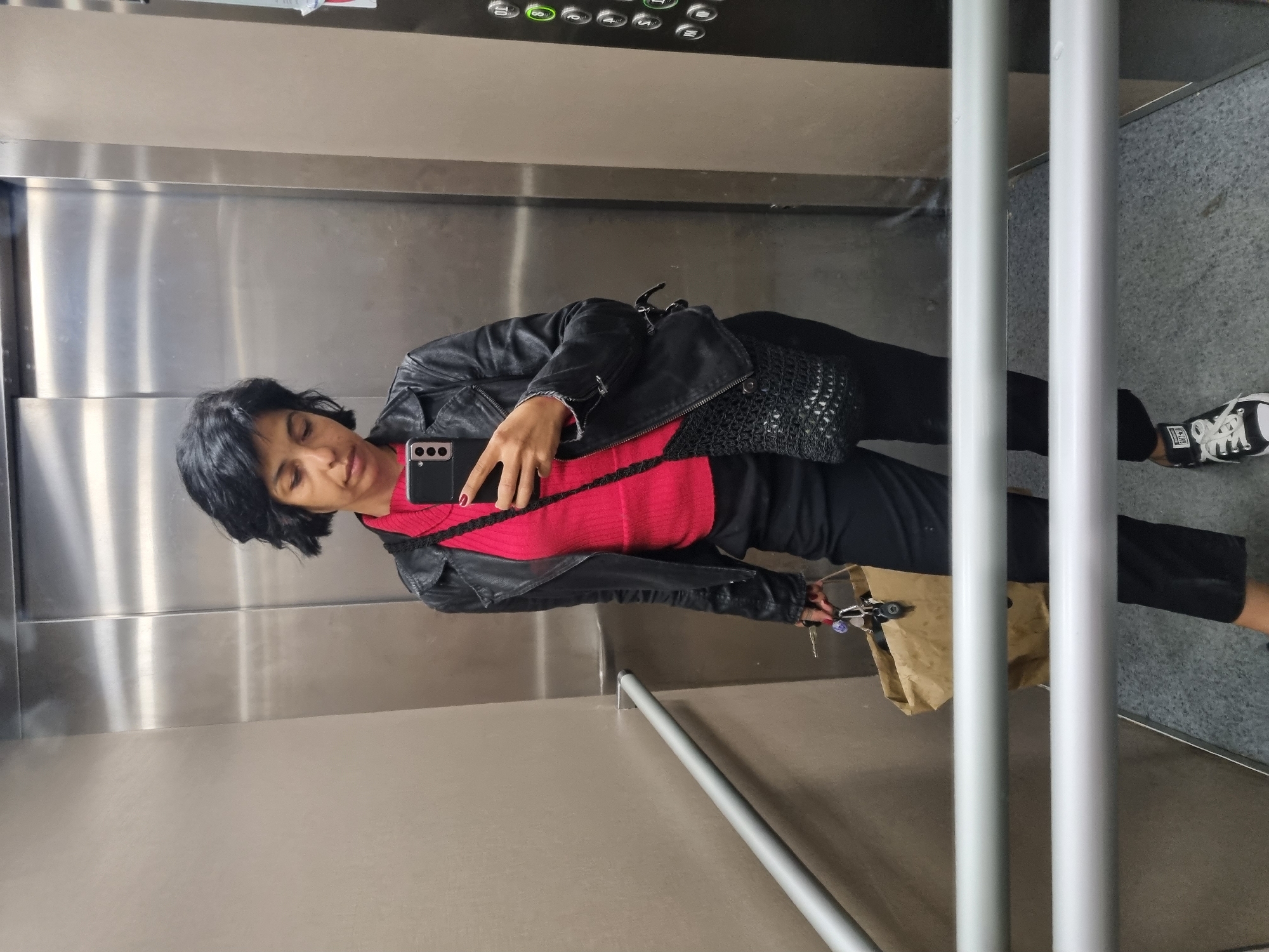 A woman takes a selfie in an elevator. She is wearing a black leather jacket over a red shirt, black pants, and black and white sneakers. She is holding a cell phone in her right hand. A brown paper bag and keys are in her left hand. The elevator has stainless steel walls and a gray floor. The buttons are in the top corner, near the ceiling. There are two vertical white handrails. The scene appears to be in an elevator, but there is no specific information to determine that it is in Plovdiv, Bulgaria.