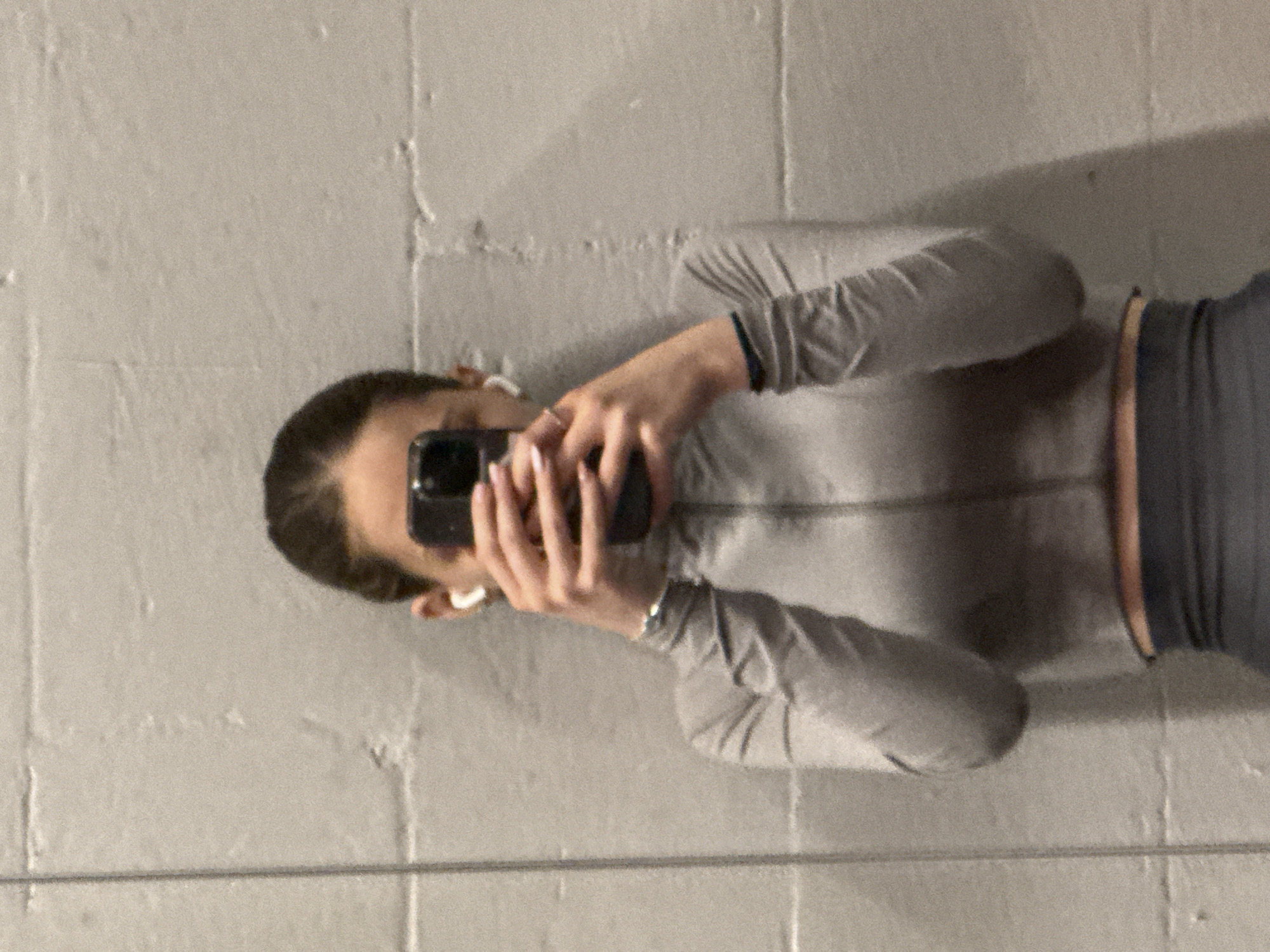 A woman is holding up a cell phone, obscuring her face, as if taking a selfie in front of a grey, textured wall. She is wearing a light grey, long-sleeved zippered jacket and dark grey high-waisted leggings, with a thin copper-colored strip of skin visible between the jacket and leggings. She has white earbuds in both ears. The wall behind her appears to be made of concrete blocks, possibly in an indoor space like a garage or basement. The location in Sofia, Bulgaria, cannot be determined from the image alone.