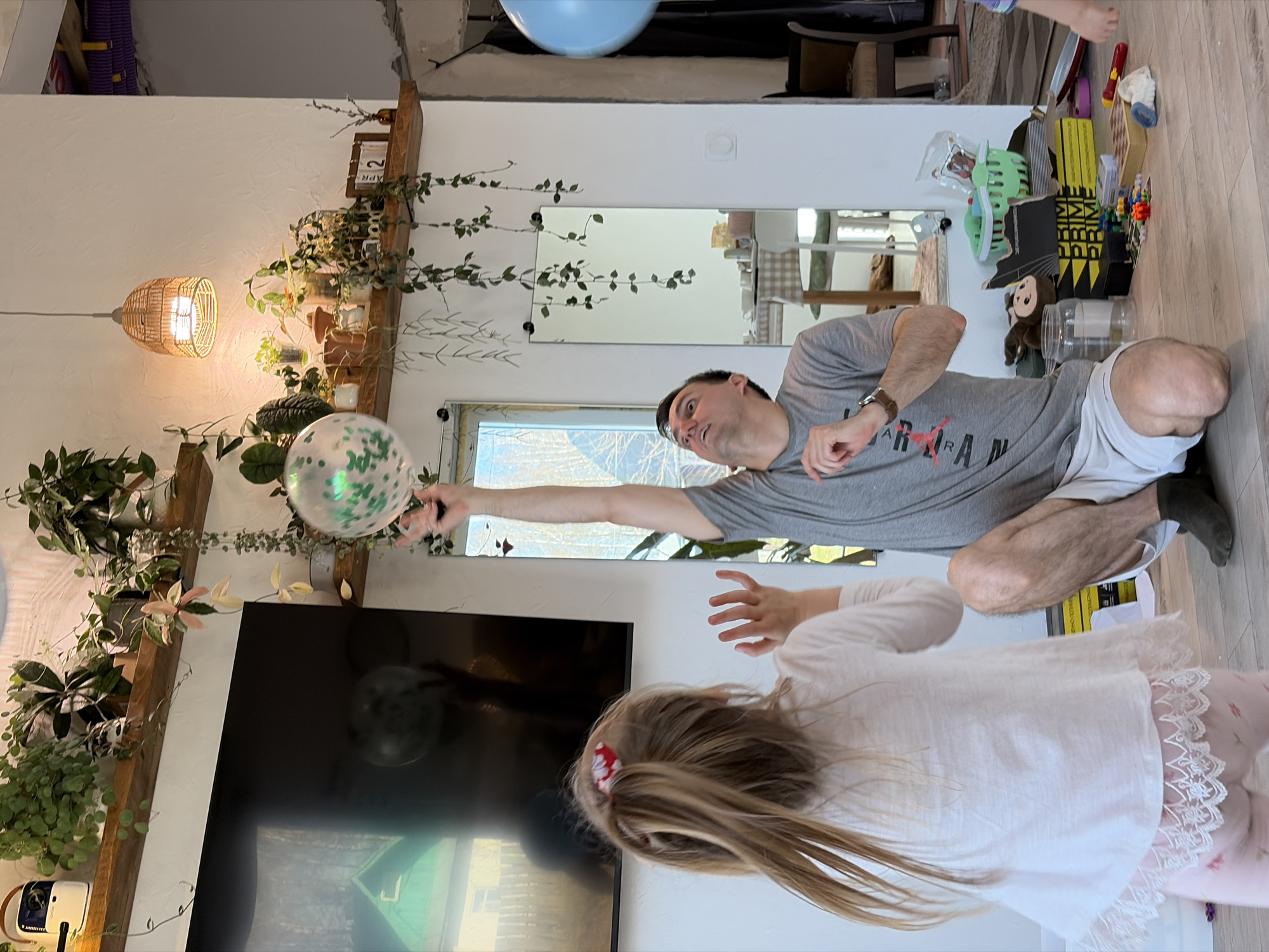 The image shows a man and a young girl indoors. The man, wearing a grey t-shirt and shorts, is reaching toward a balloon decorated with green confetti. The girl, dressed in a white top with lace detailing, seems to be reaching for the balloon as well. A television is visible in the background, along with shelves holding plants and other decorative items. There's a mirror on the wall and various objects, including toys and containers, scattered on the floor. The location is a domestic interior. There is no indication of the city of Malyi Bukor, Russia.